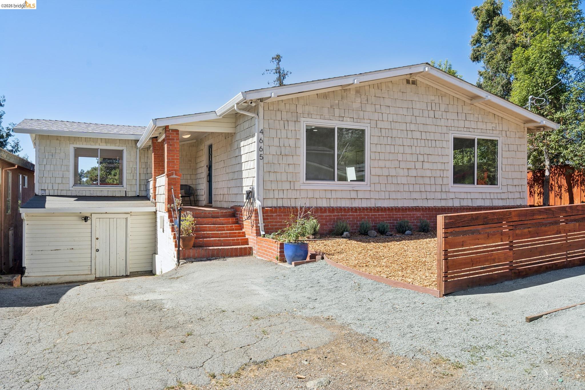 Light-shingled home with brick steps, gravel driveway, and wooden front fence.