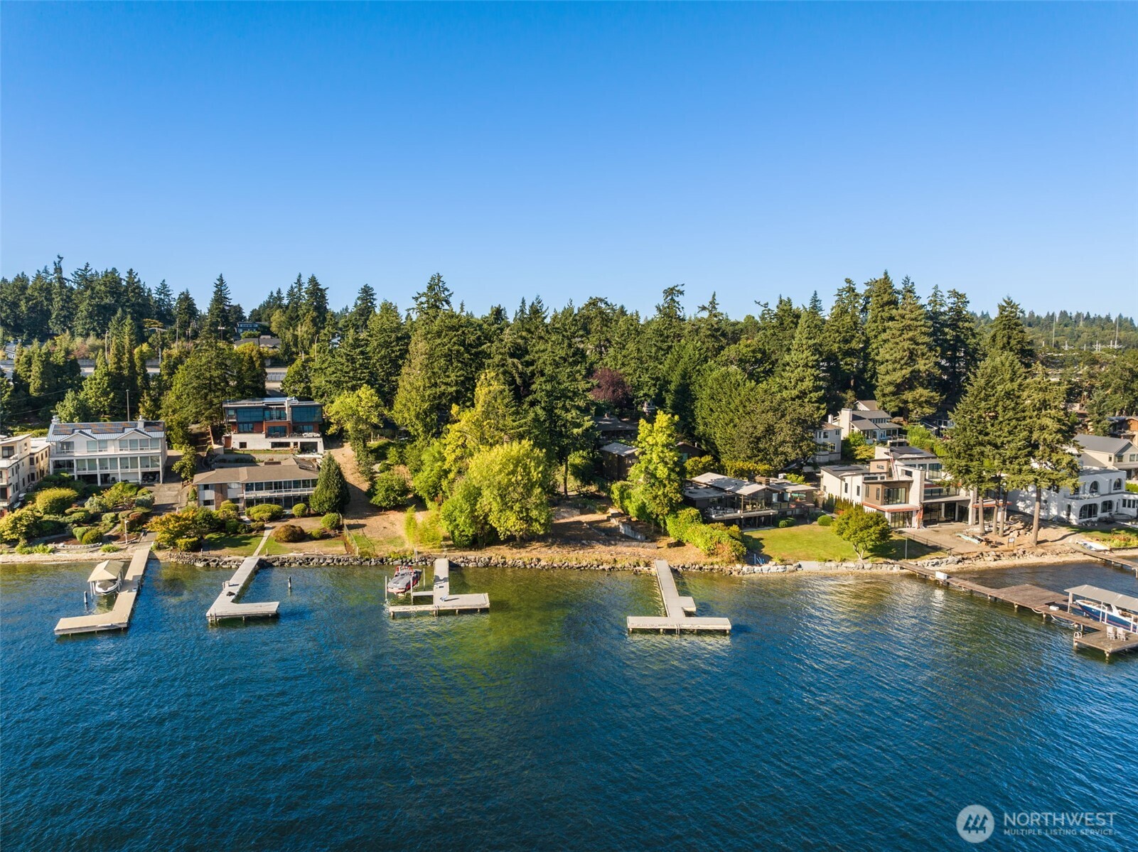 Aerial view of wooded lakeshore homes, docks, and waterfront on a sunny day.