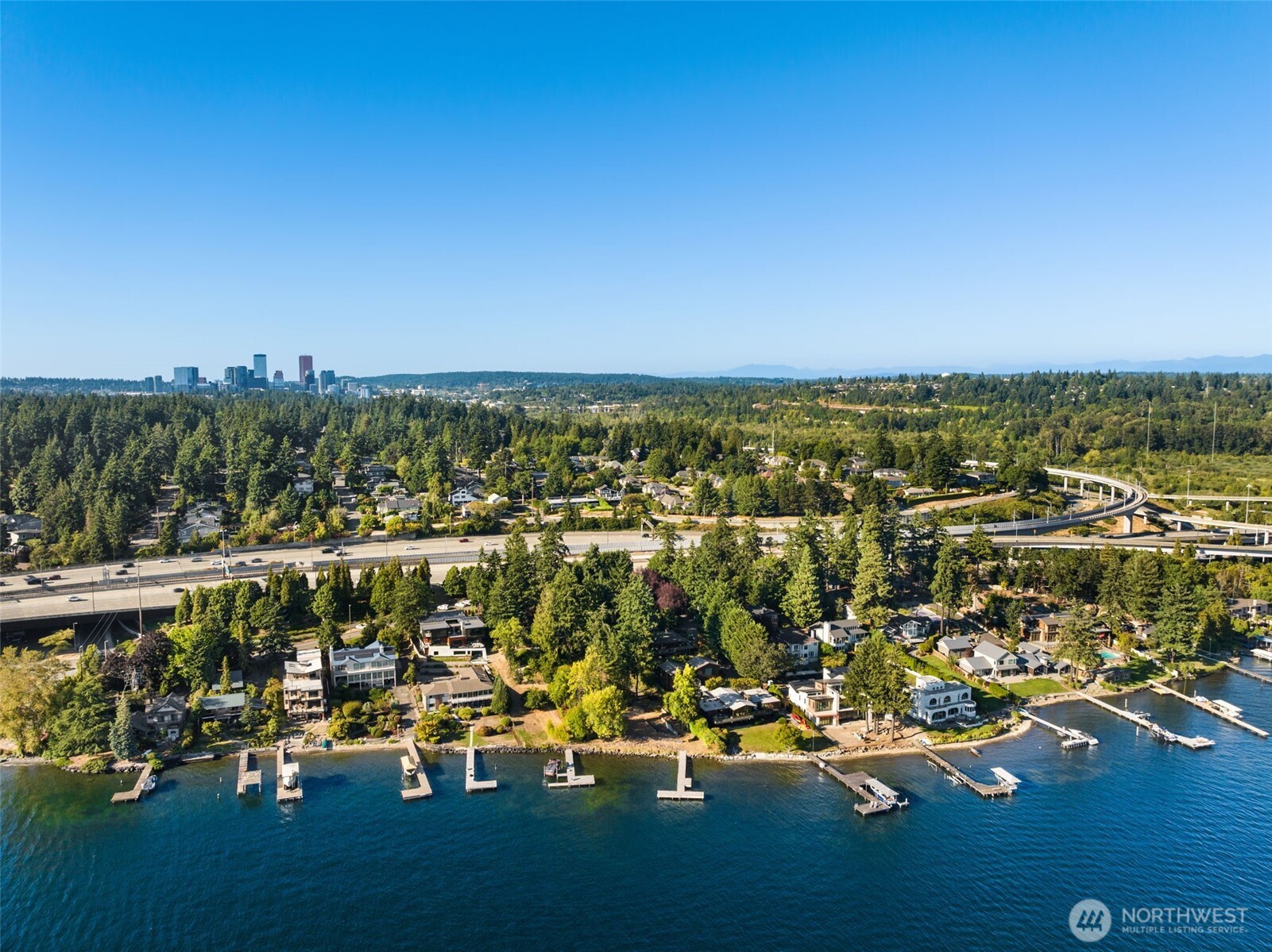 Aerial shoreline homes and docks on Lake Washington, with a distant city skyline and curving highway.