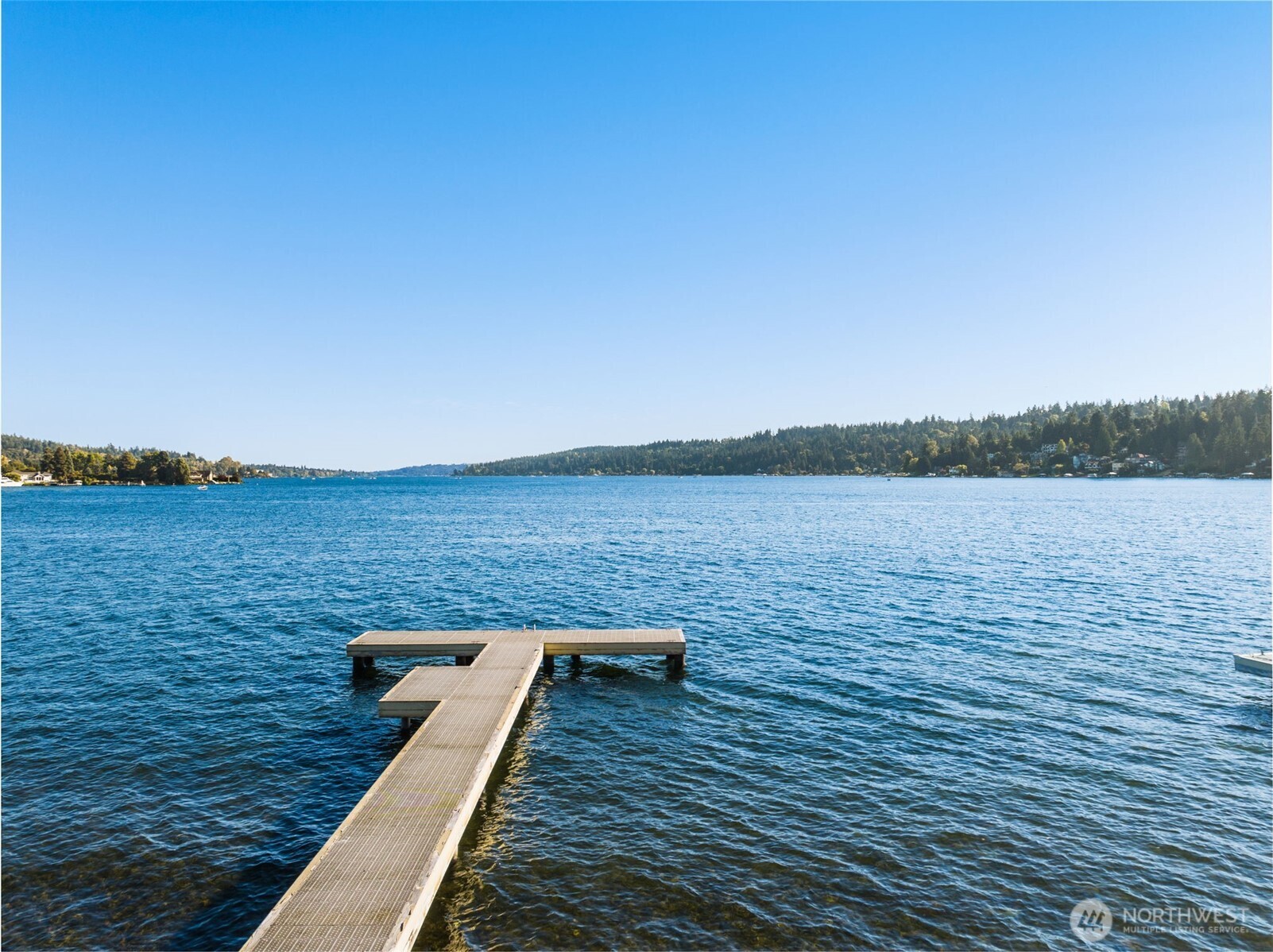 T-shaped dock extends into a blue lake, with forested shorelines across the water.