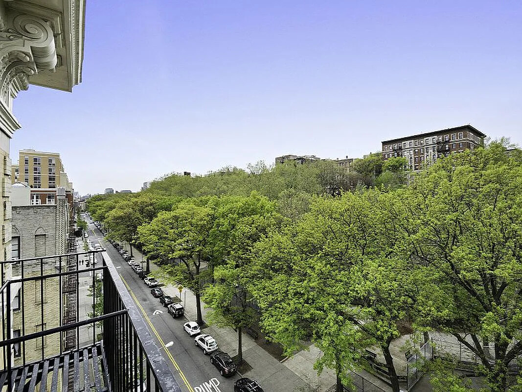 Manhattan street view from a balcony, with leafy trees, parked cars, and city buildings.