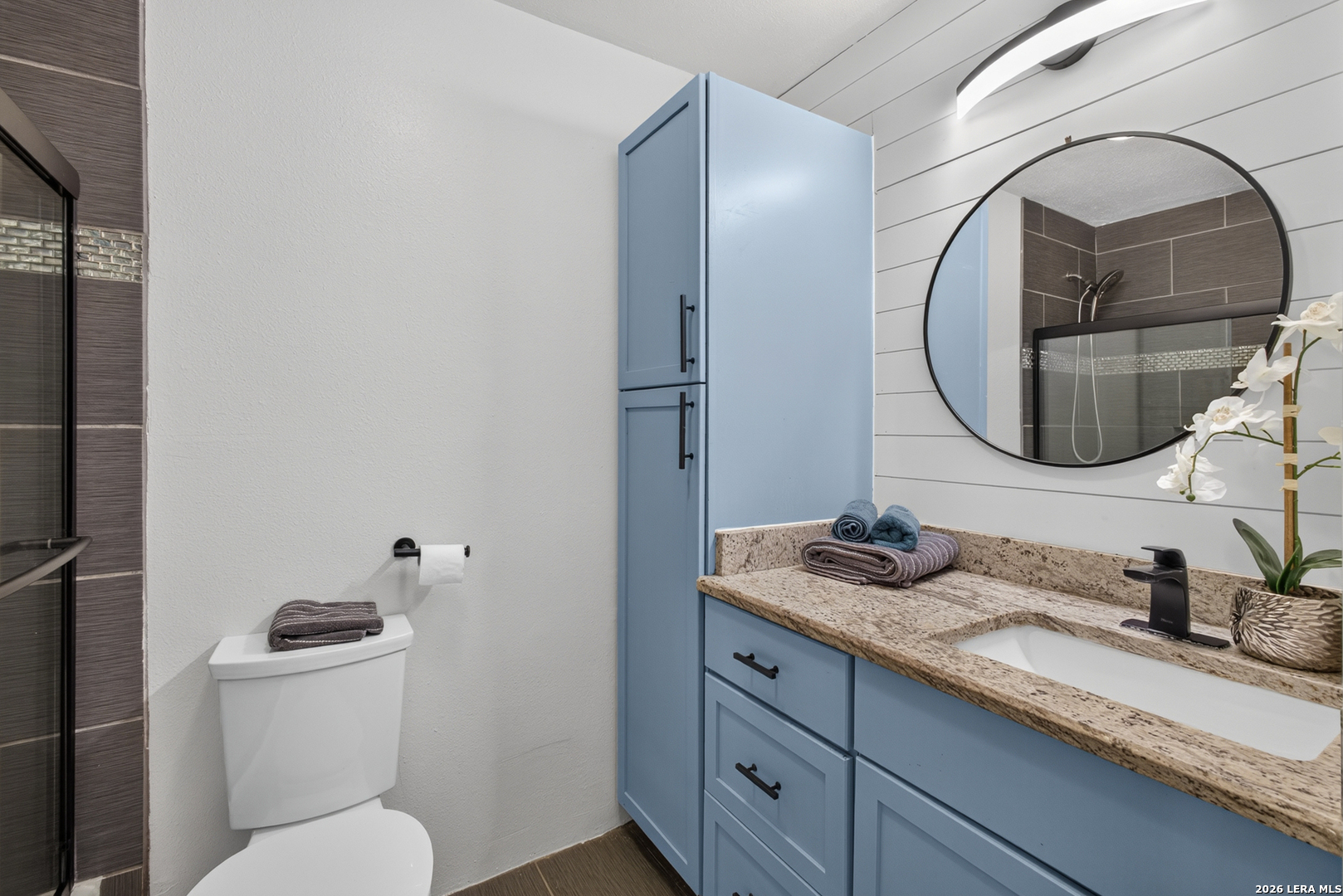 Bathroom with blue vanity, granite countertop, round mirror, and glass-enclosed shower.