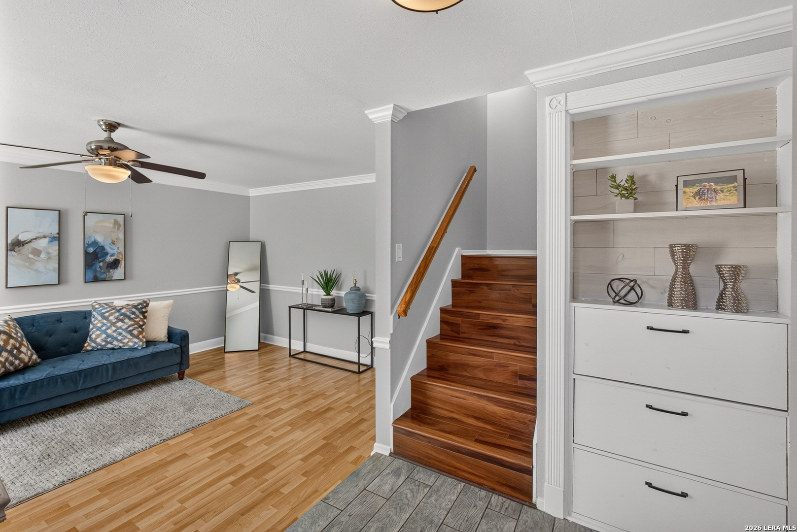 Staged living room with a blue sofa, hardwood floors, built-in shelving, and a staircase.