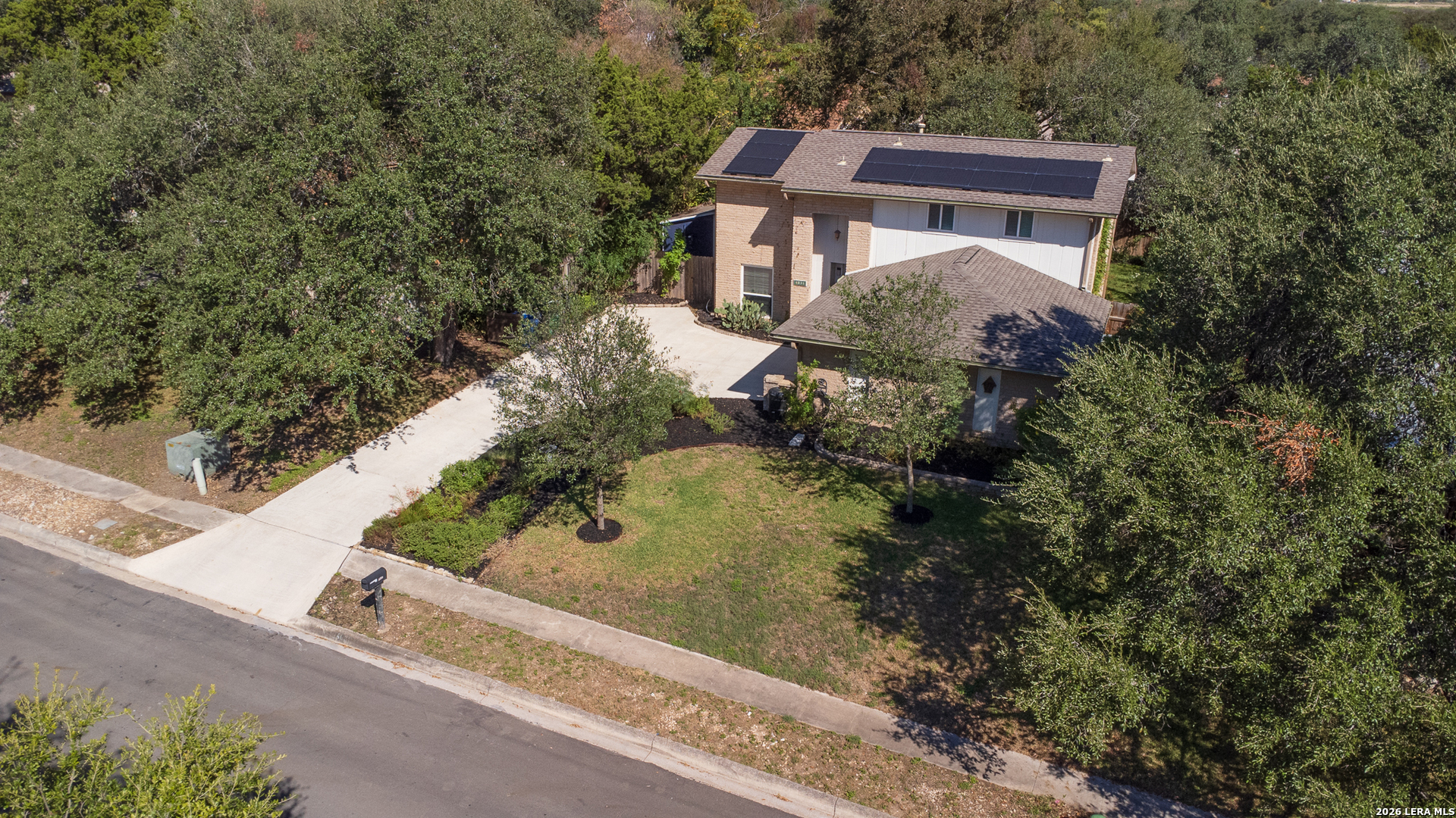 Aerial view of a two-story brick home with rooftop solar panels, a broad driveway, and mature trees.