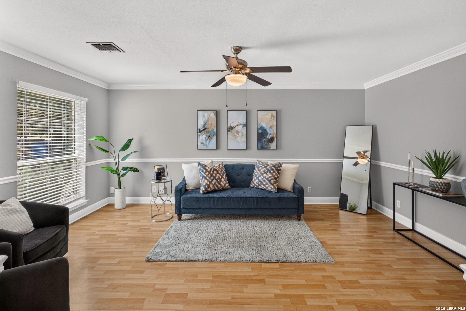 Staged living room with a blue sofa, gray walls, wood flooring, and a ceiling fan.