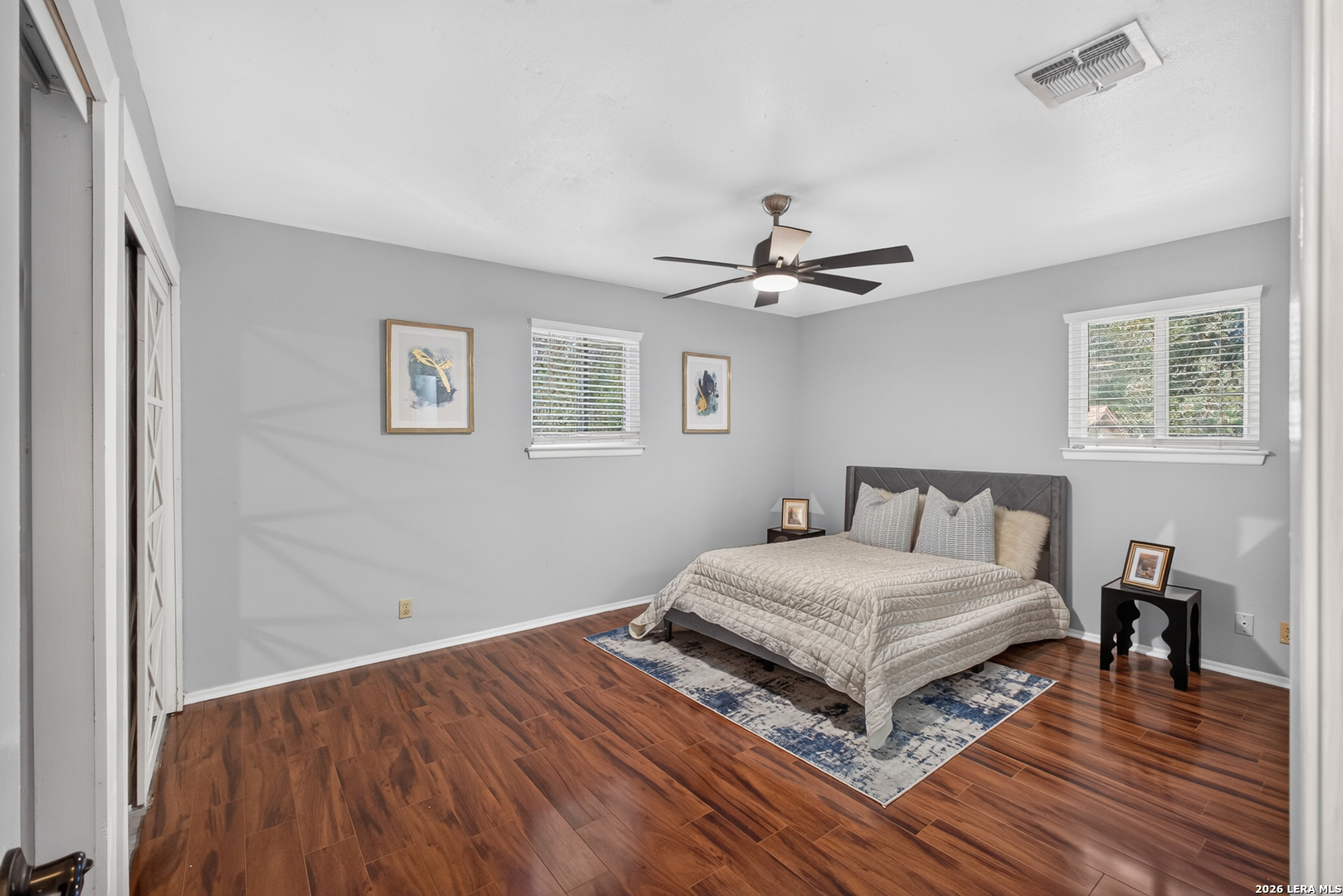 Bedroom with a gray upholstered bed, wood-look flooring, and two windows with blinds.