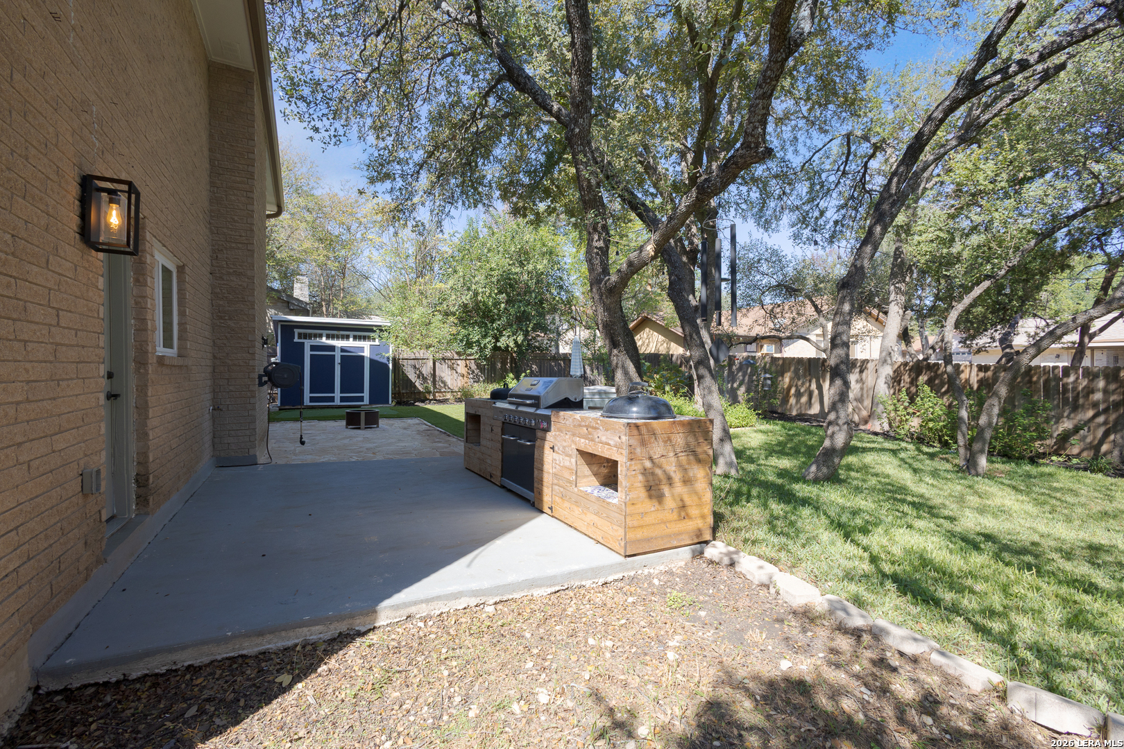Backyard patio with a built-in outdoor grill, mature trees, and a blue storage shed.