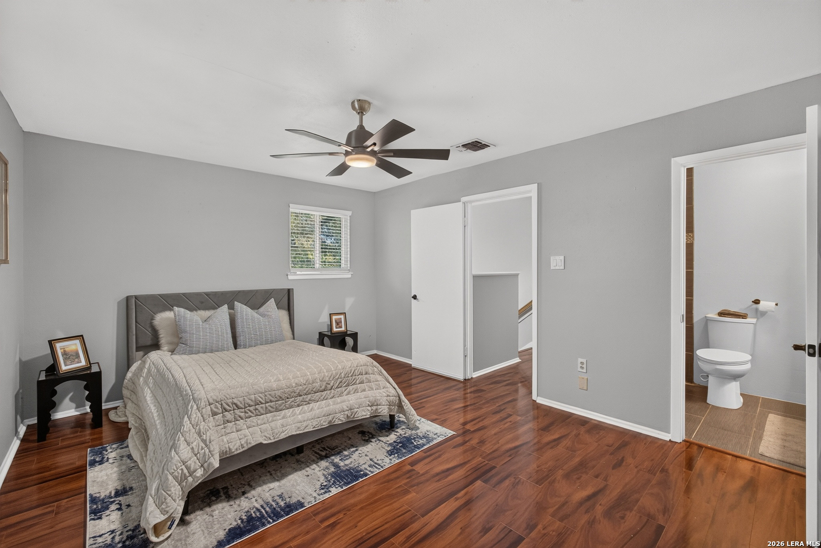 Staged bedroom with a quilted bed, ceiling fan, wood floors, and an adjoining bathroom.