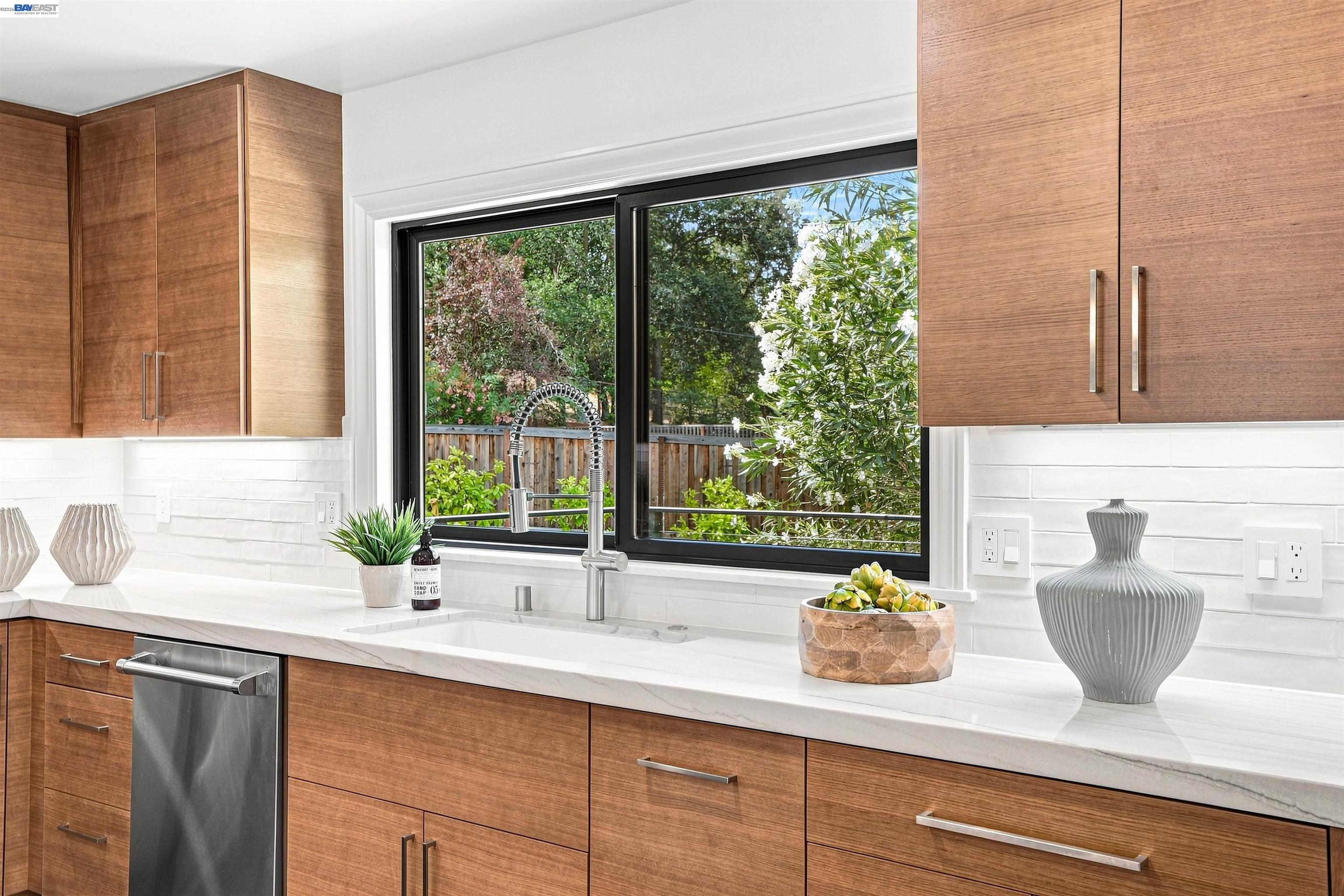 Kitchen with warm wood cabinets, white counters, and a large window overlooking a leafy yard.