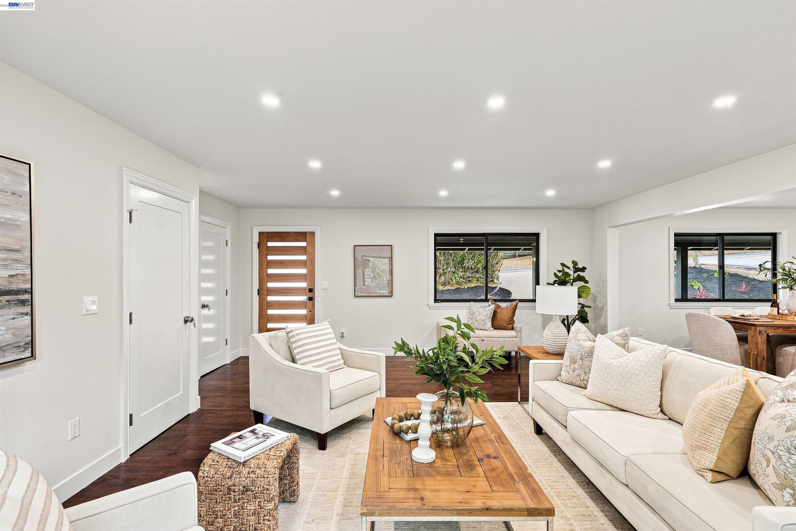 Staged living room with white seating, a wooden coffee table, and windows overlooking landscaping.