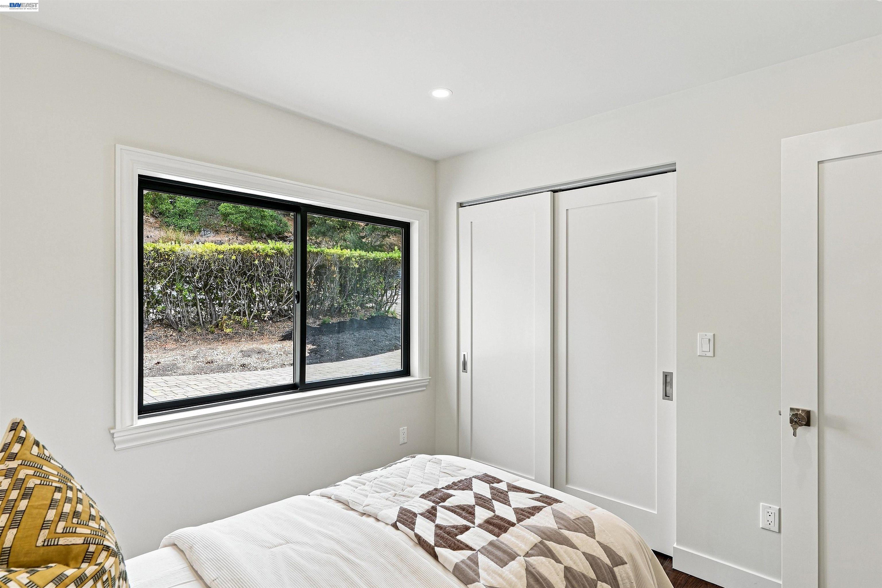 Furnished bedroom with a black-framed window overlooking hedges and white sliding closet doors.