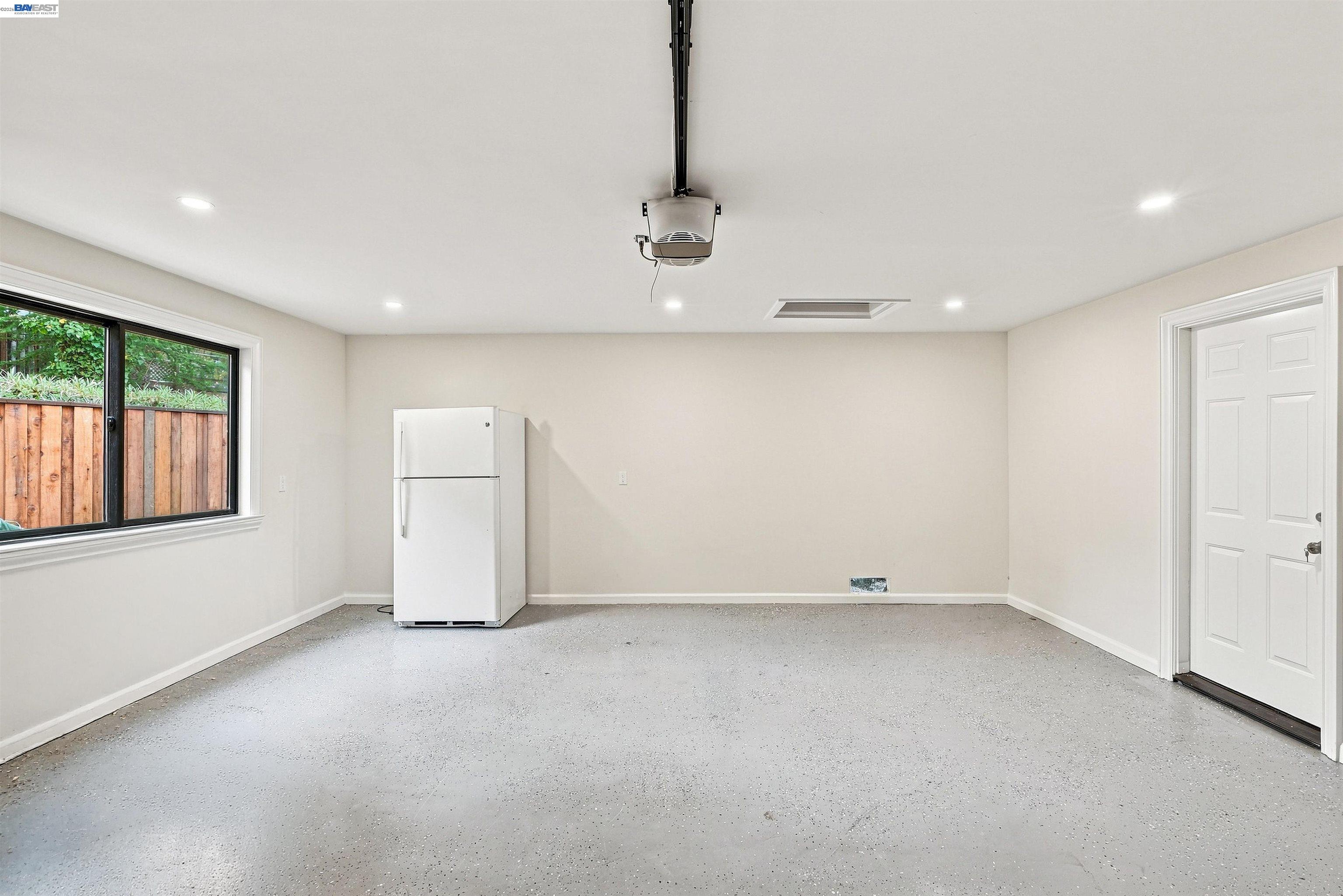 Vacant garage with a white refrigerator, speckled concrete floor, and window overlooking a wooden fence.