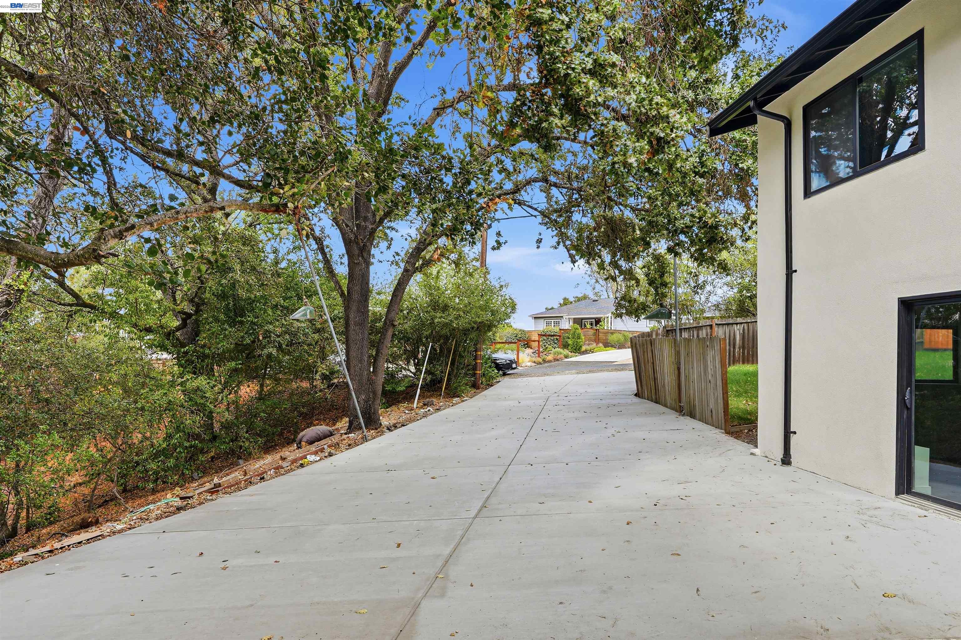 Concrete driveway beside a light-colored home, shaded by mature trees and bordered by a wooden fence.