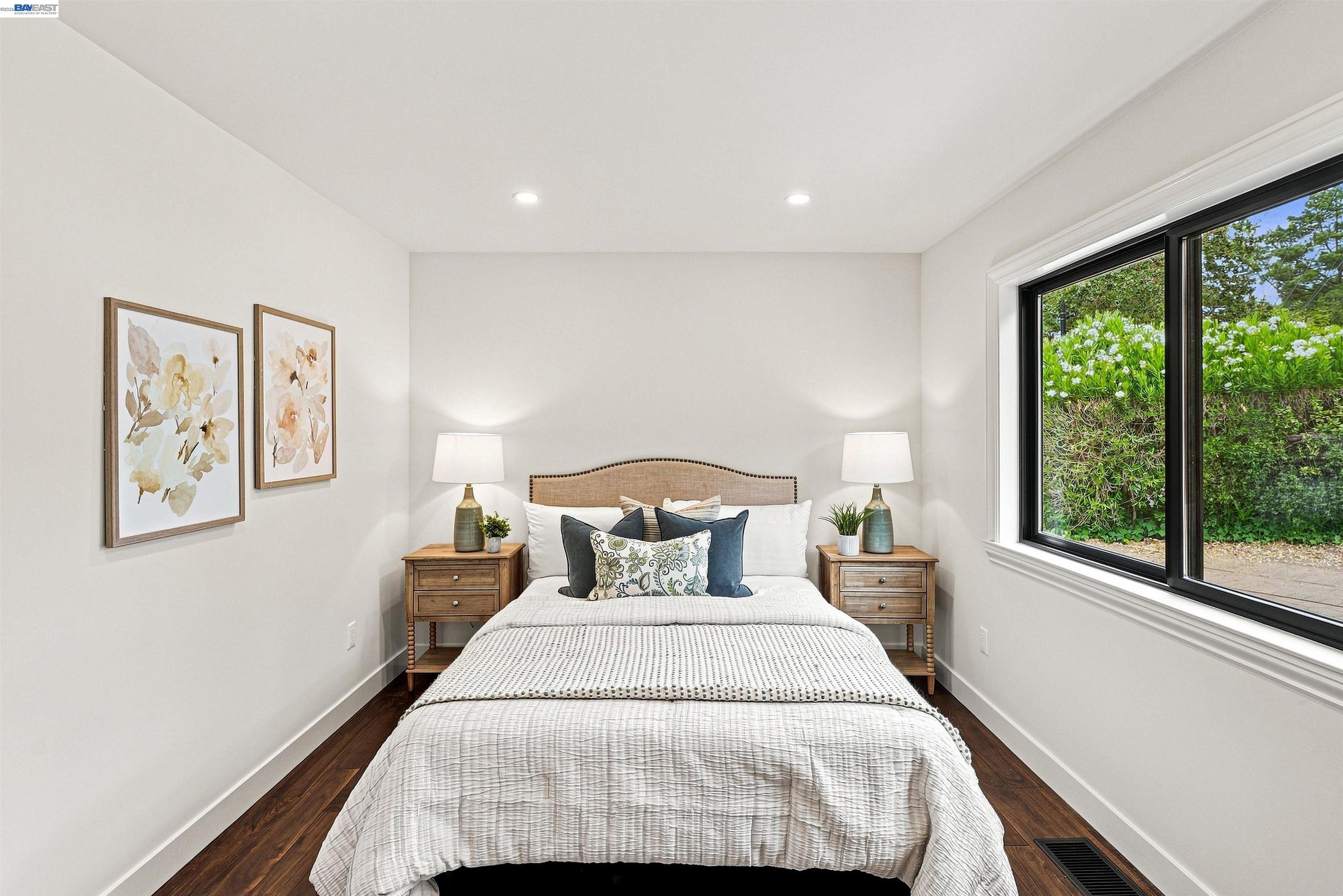 Staged bedroom with a centered bed, matching nightstands, floral prints, and a large window overlooking greenery.