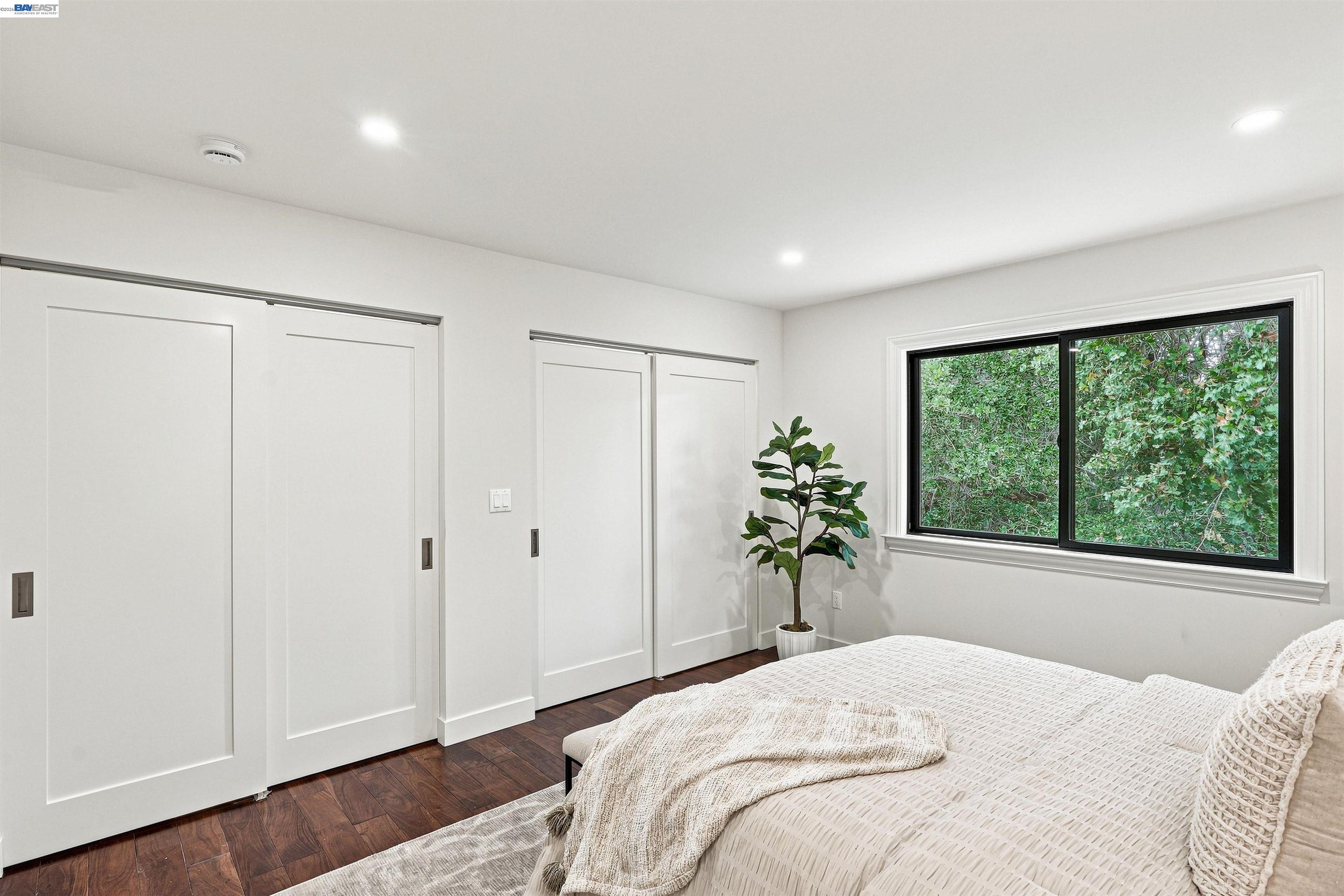Staged bedroom with white sliding closets, dark hardwood floors, and a window overlooking leafy trees.