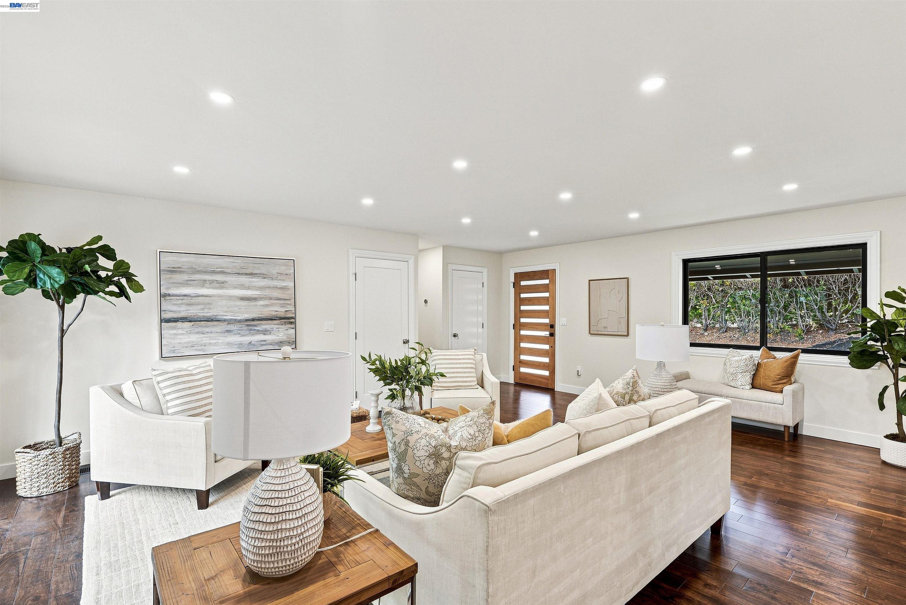 Staged living room with cream seating, dark wood floors, and a large window overlooking shrubs.