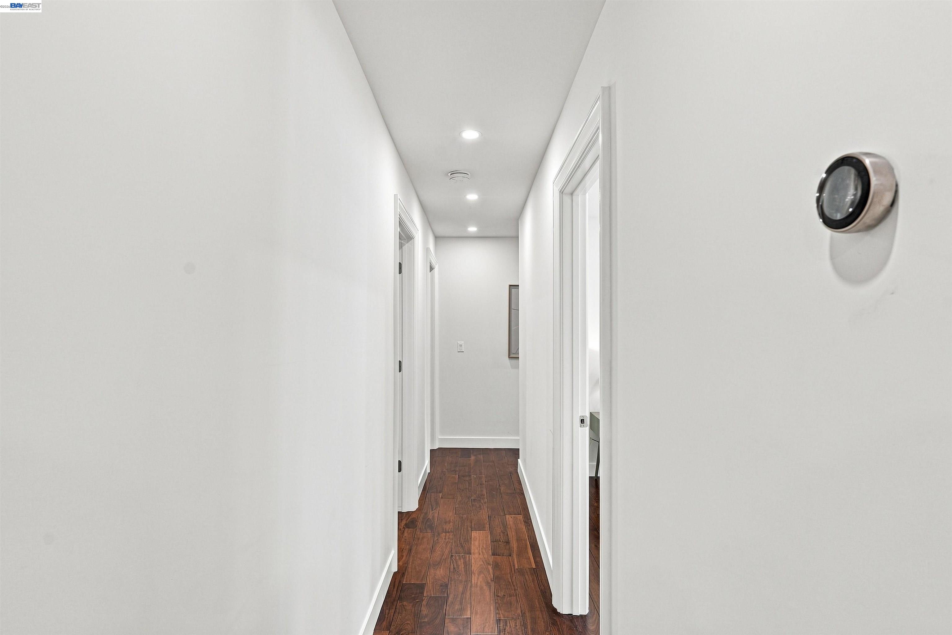 Bright white hallway with dark wood flooring, recessed lights, and a round wall thermostat.