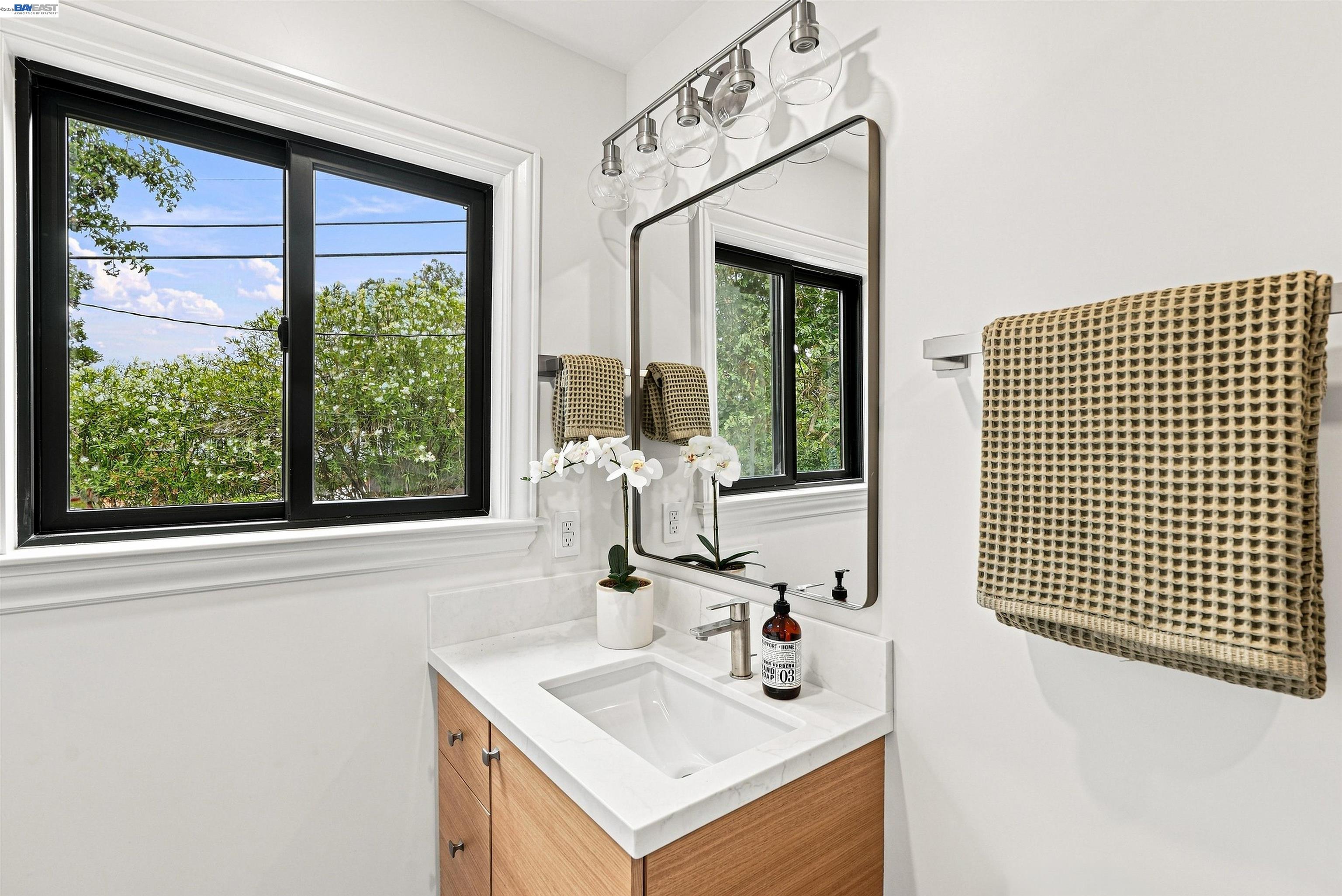 Bathroom vanity with a white sink, wood cabinetry, large mirror, and black-trimmed windows.