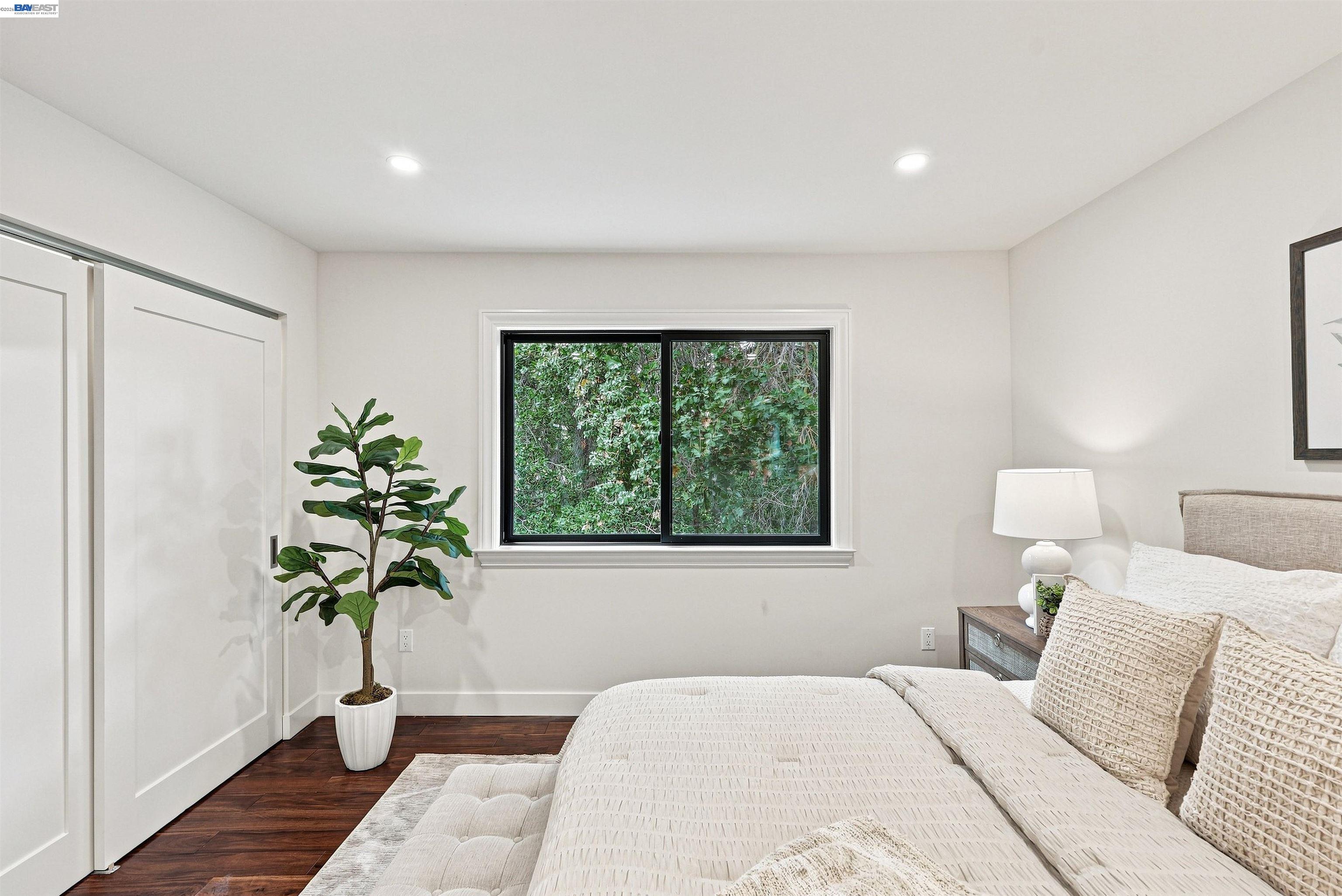 Staged bedroom with white bedding, dark wood floors, and a window overlooking greenery.
