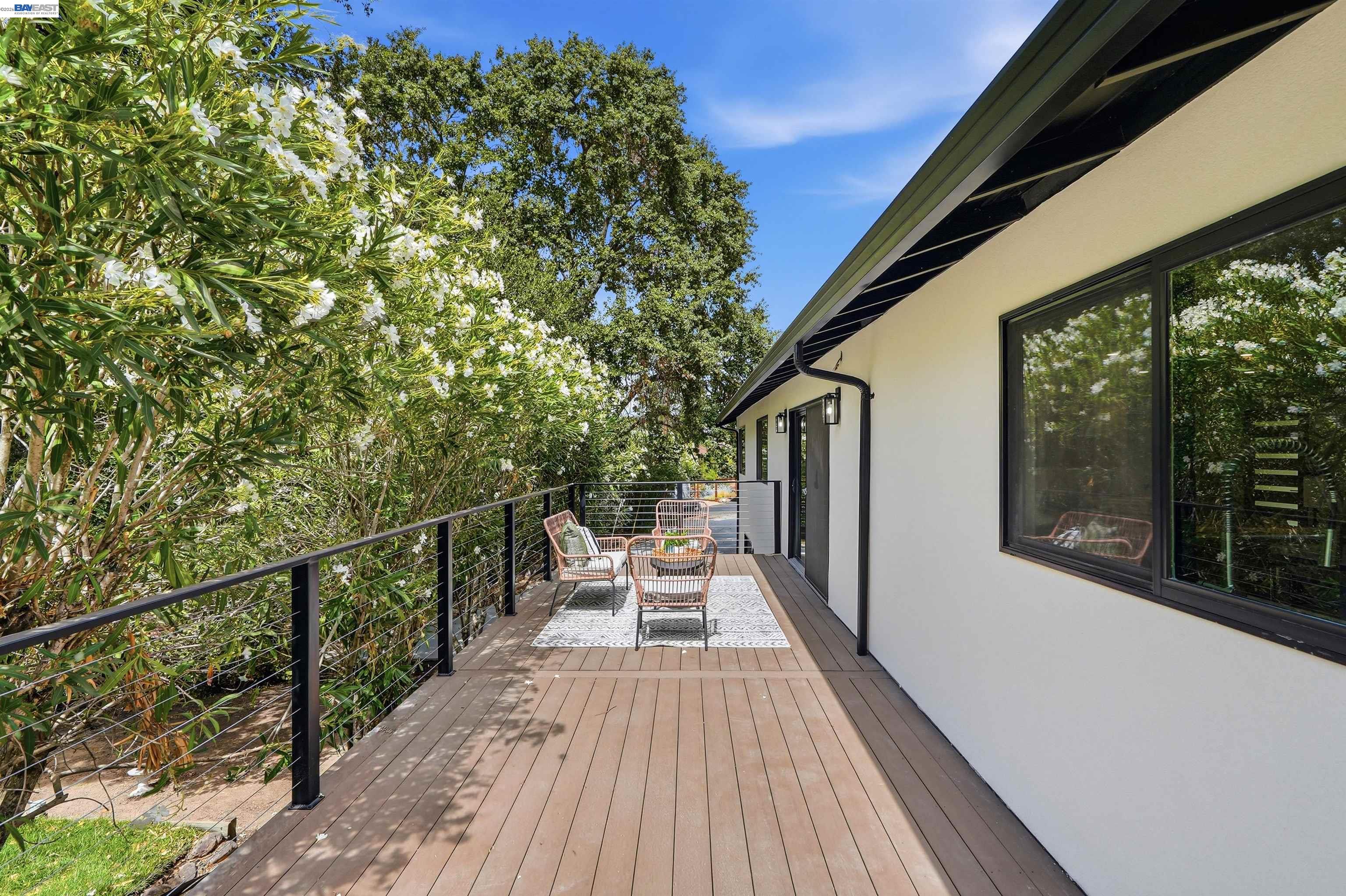 Furnished wooden deck beside a modern home, bordered by flowering greenery and black cable railings.