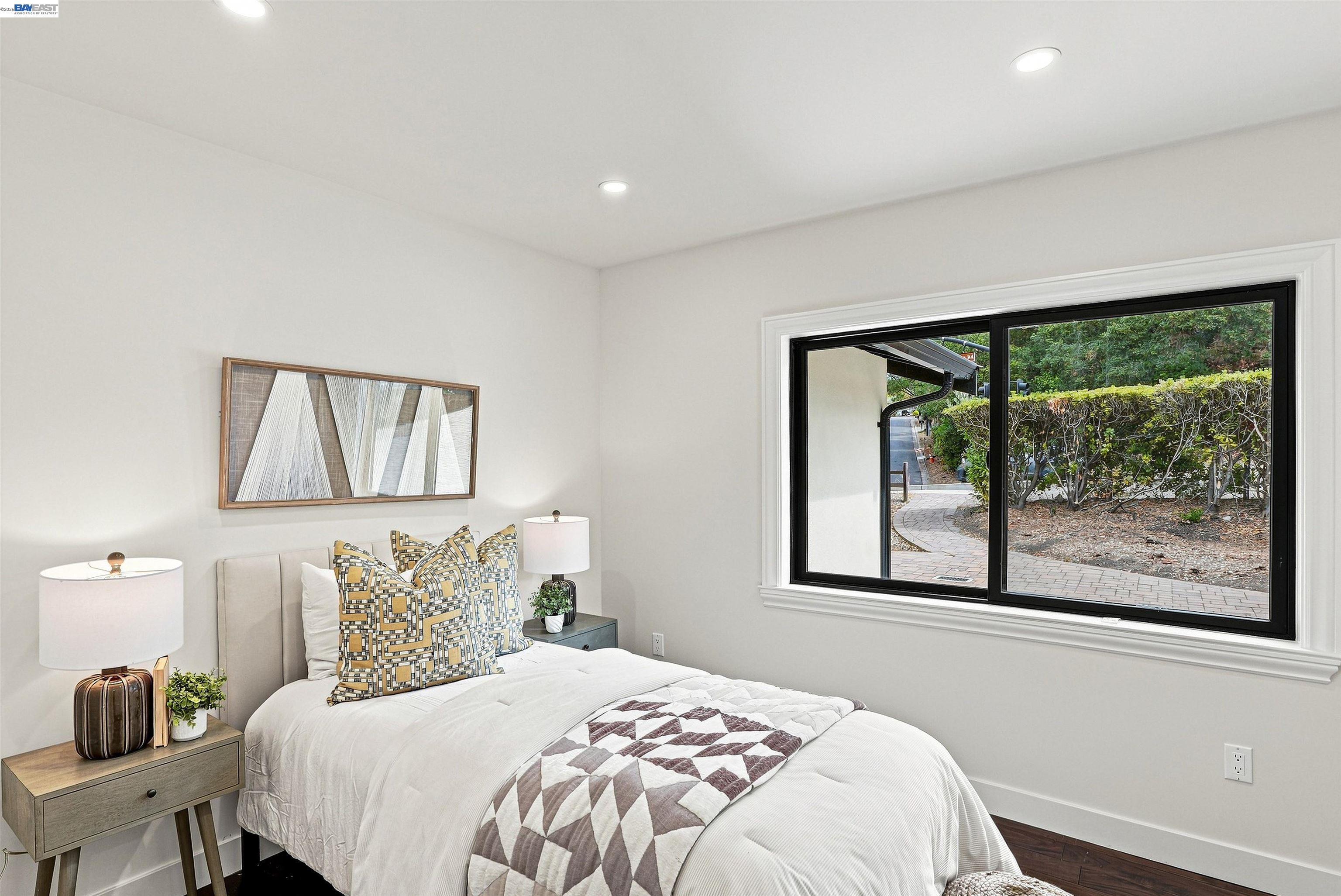 Staged bedroom with a white bed and geometric pillows beside a large window overlooking a garden path.