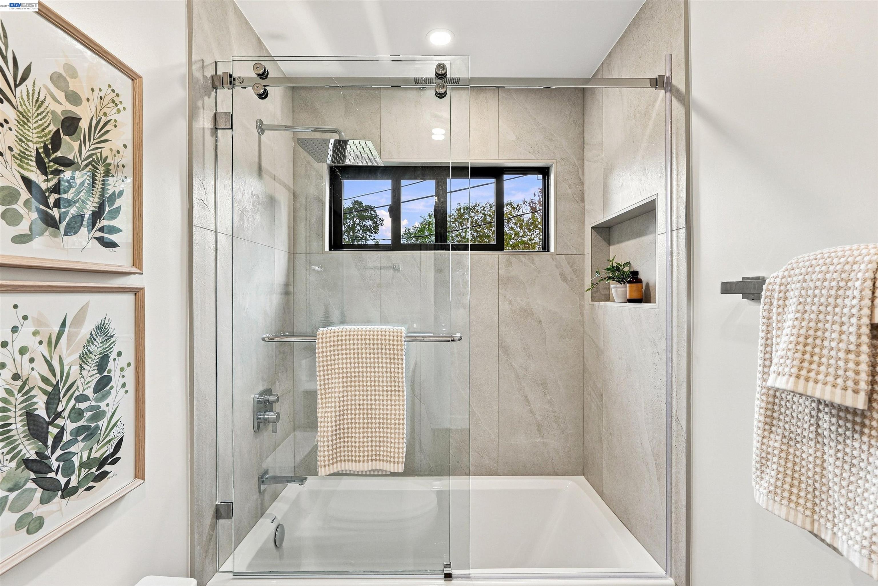 Bathroom with a tiled bathtub and glass doors beneath a black-framed window.