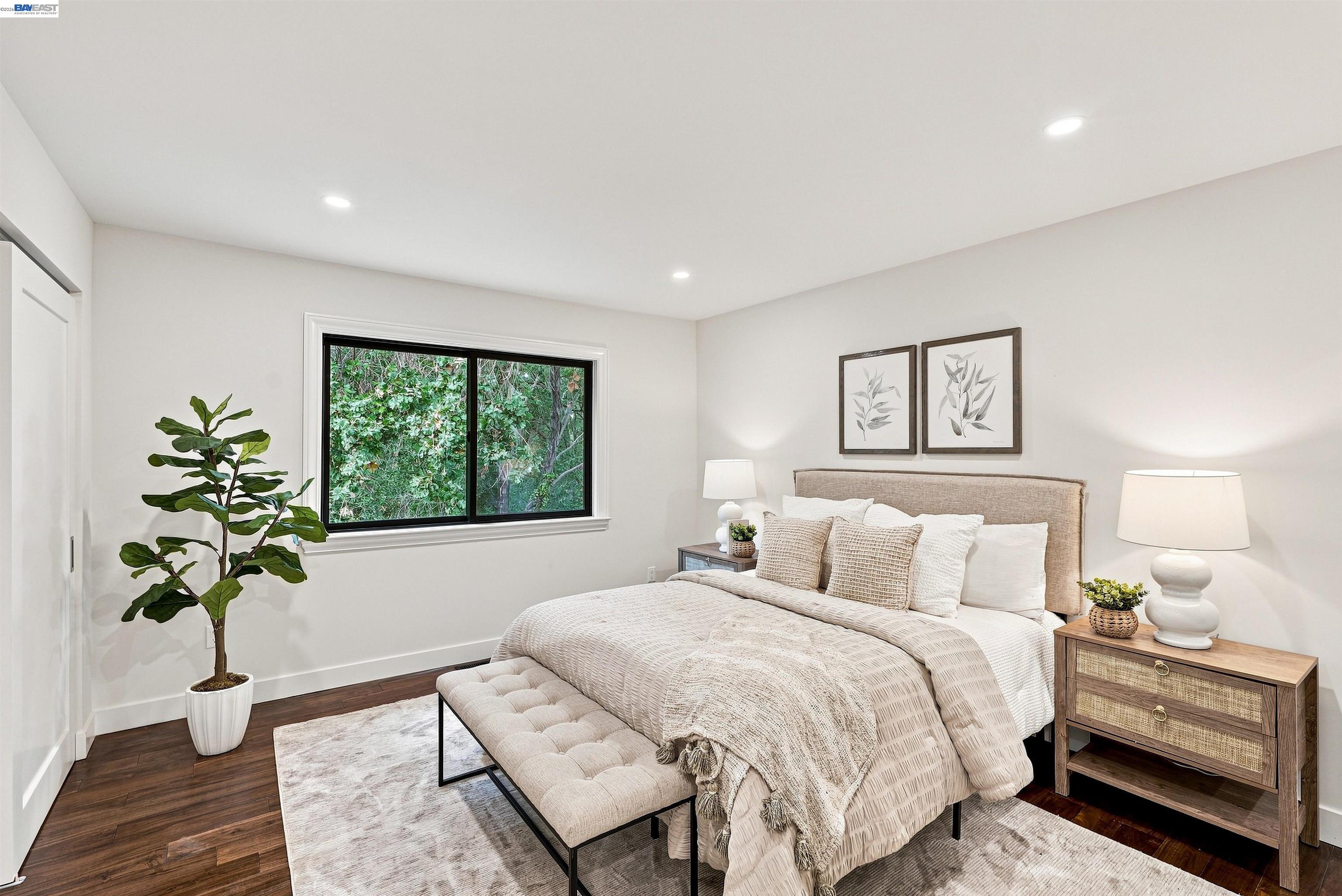 Staged bedroom with neutral bedding, a cushioned bench, and a window overlooking leafy greenery.