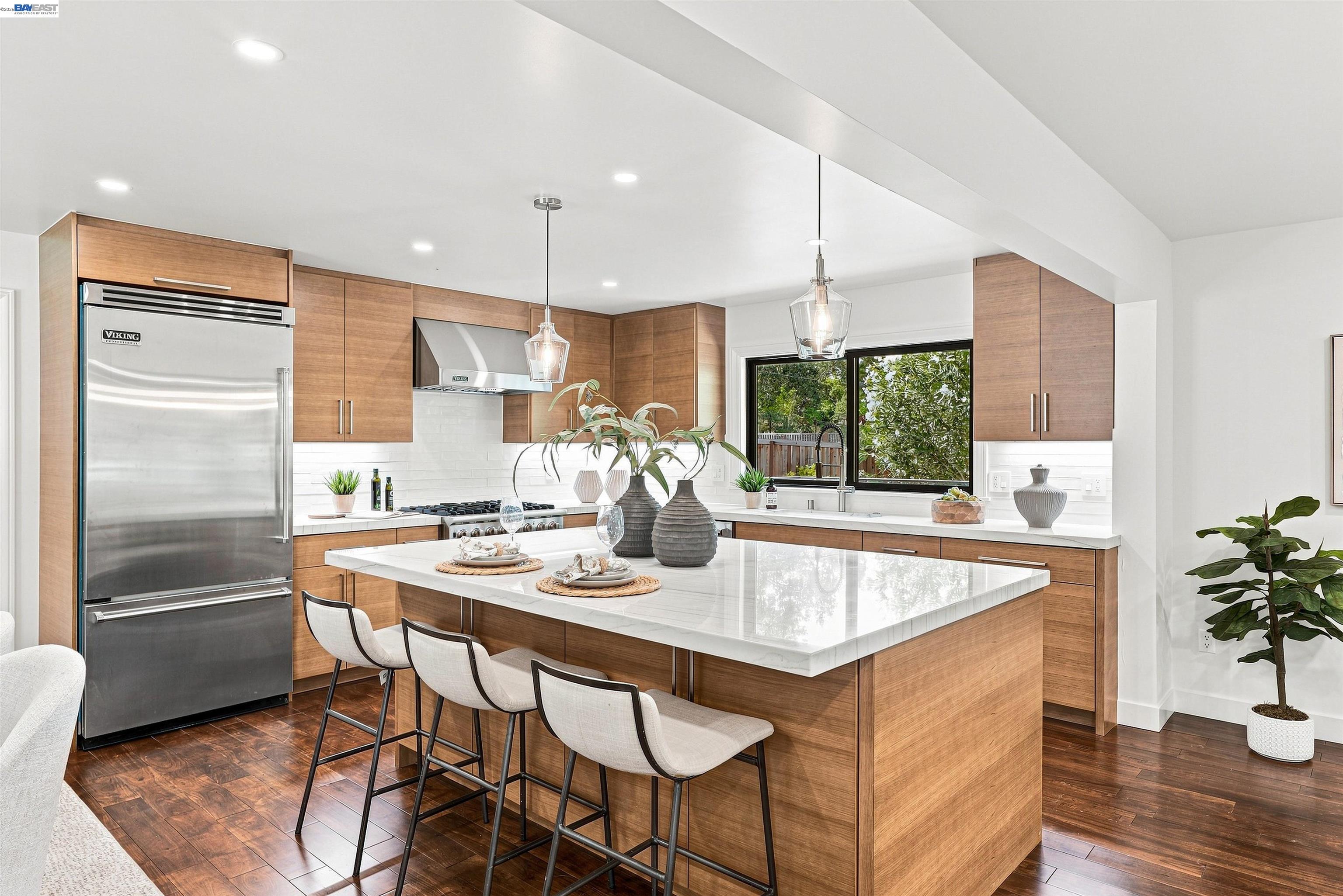 Modern kitchen with a large white island, warm wood cabinets, three bar stools, and garden views.