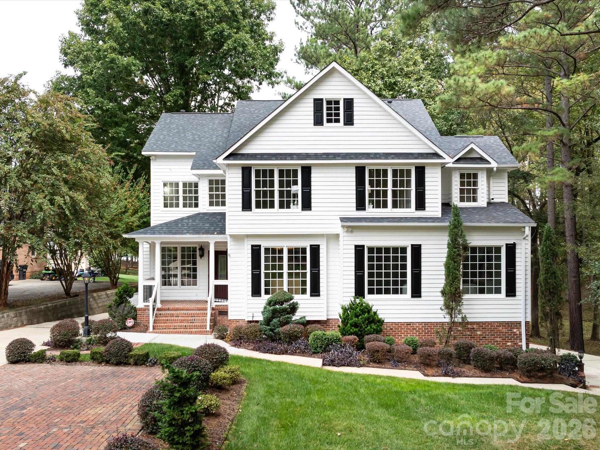 Two-story white house with black shutters, landscaped front yard, and brick driveway.