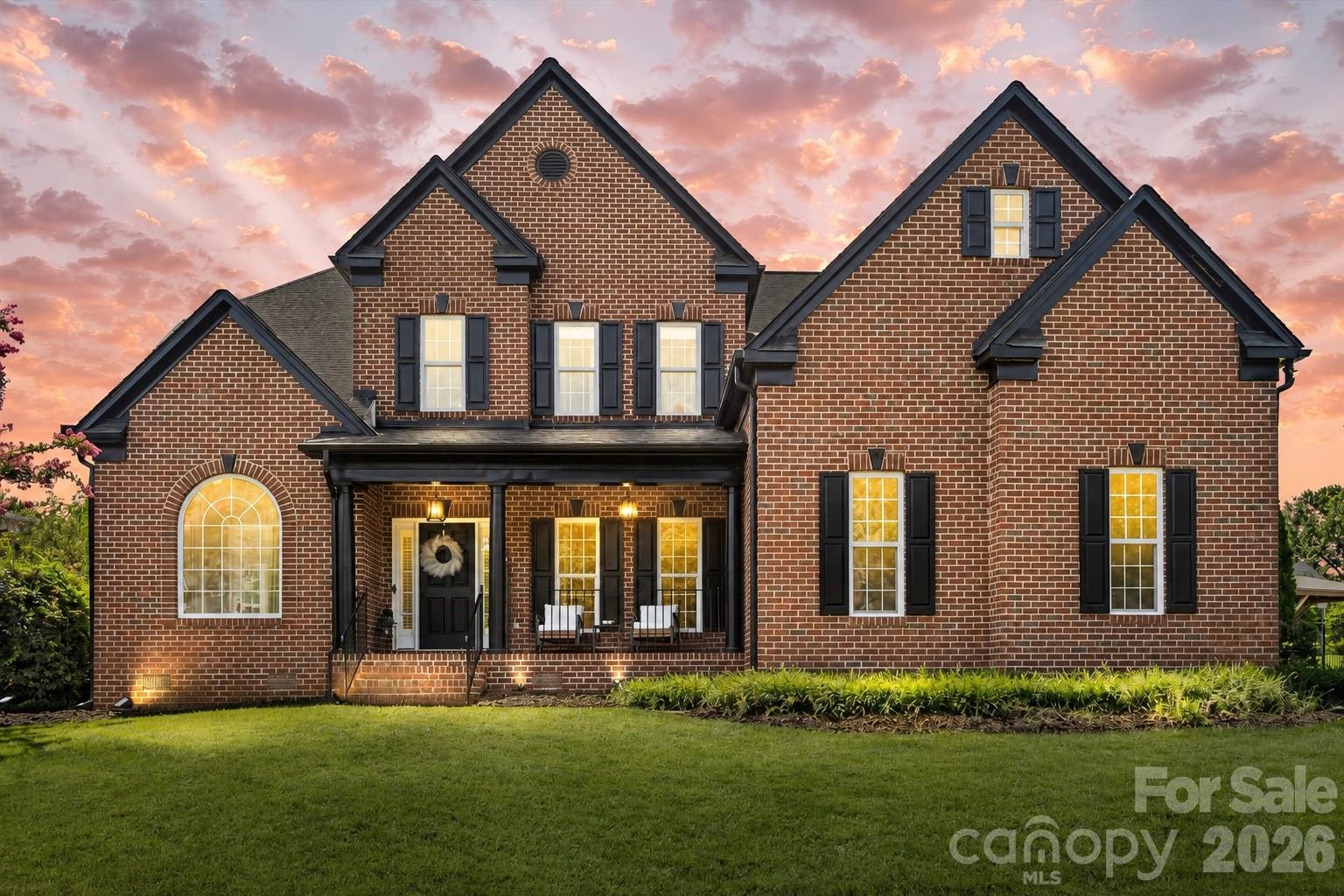 Two-story brick home with dark shutters, a covered porch, and glowing windows beneath a pink sunset sky.