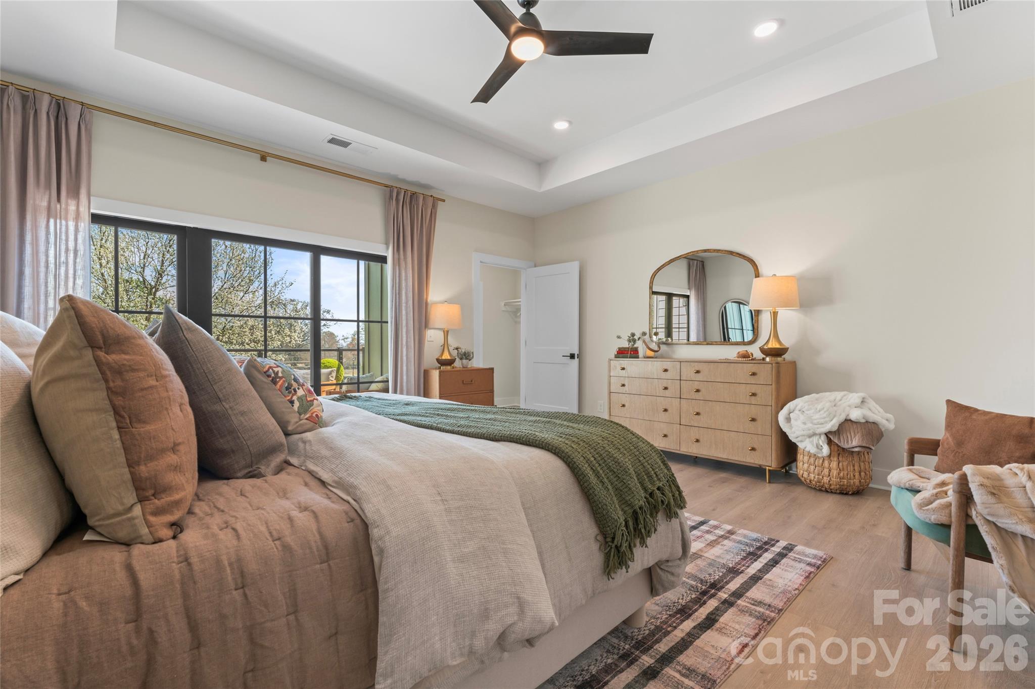 Furnished bedroom with a layered bed, green throw, tray ceiling, and large black-framed windows.