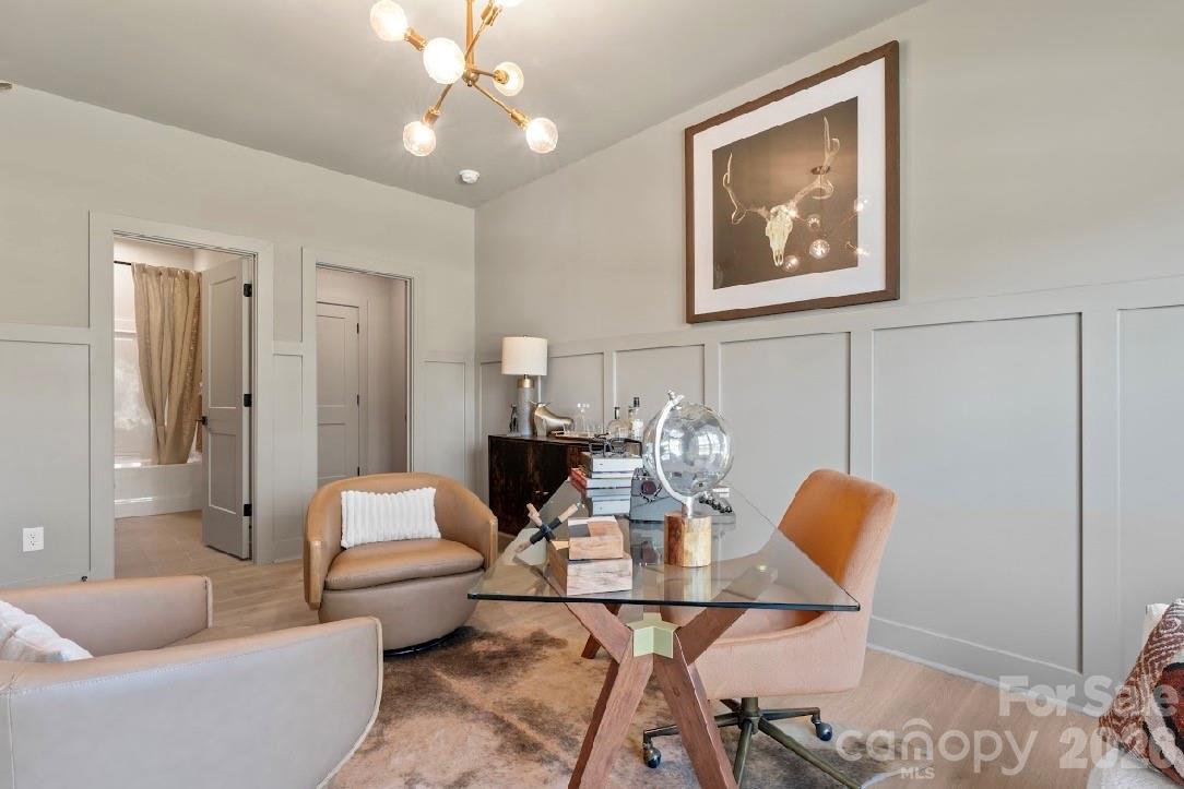 Furnished home office with a glass desk, tan chairs, paneled walls, and a modern chandelier.