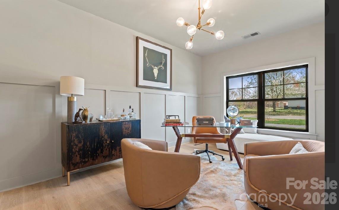 Staged home office with a glass-top desk, tan chairs, and a large black-framed window.