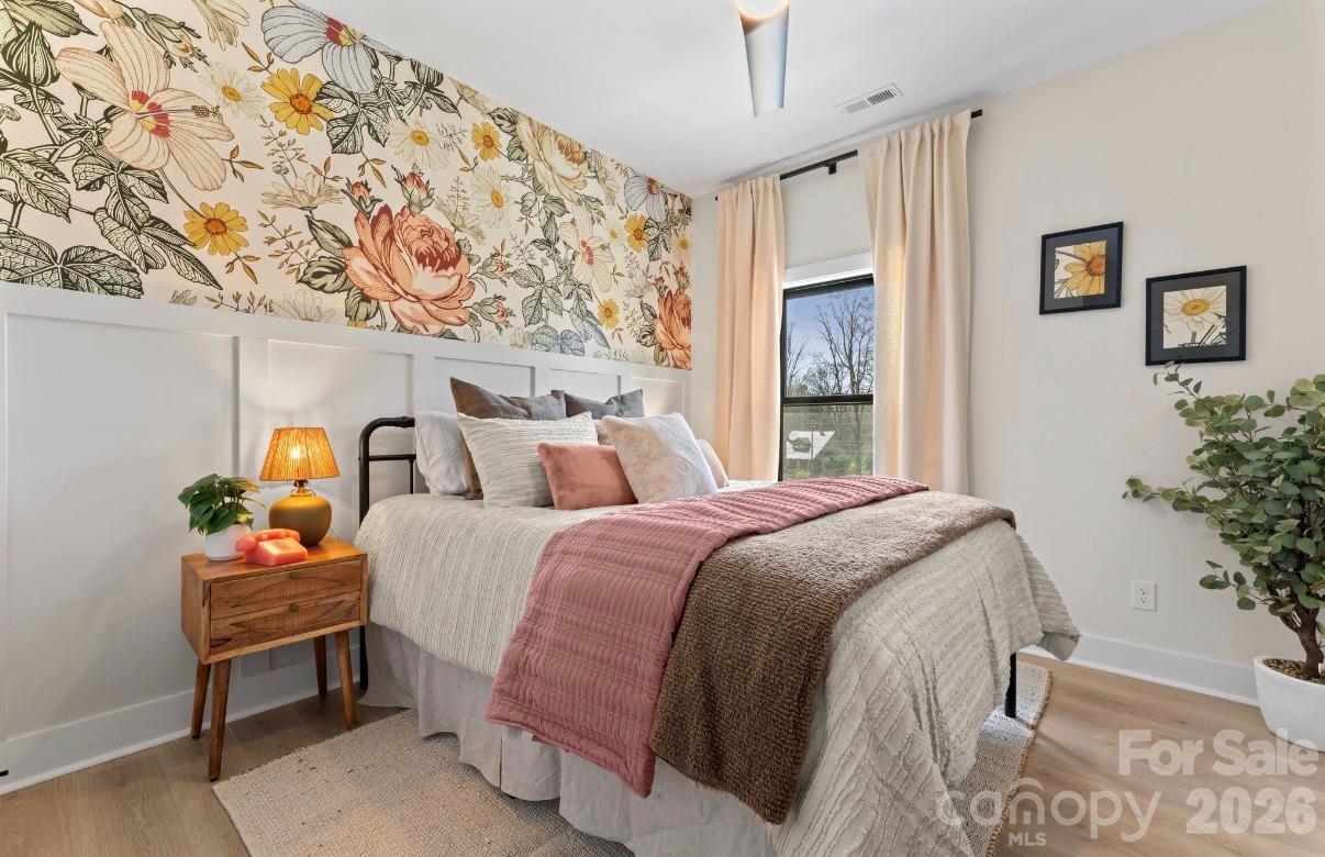 Staged bedroom with floral wallpaper, a cream bed with rose-colored bedding, and a wood bedside table.
