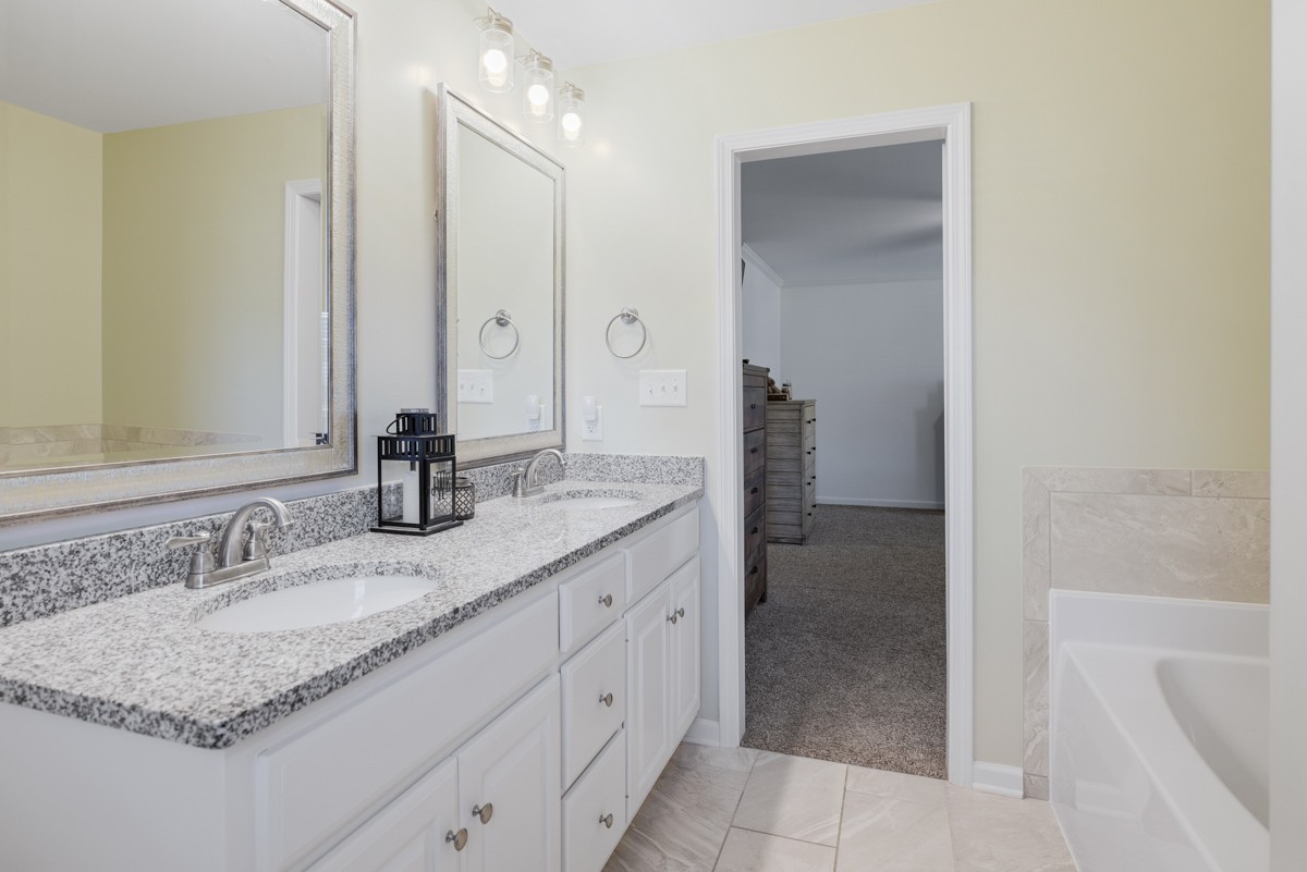 Bathroom with a double-sink granite vanity, framed mirrors, and a bathtub beside an open doorway.