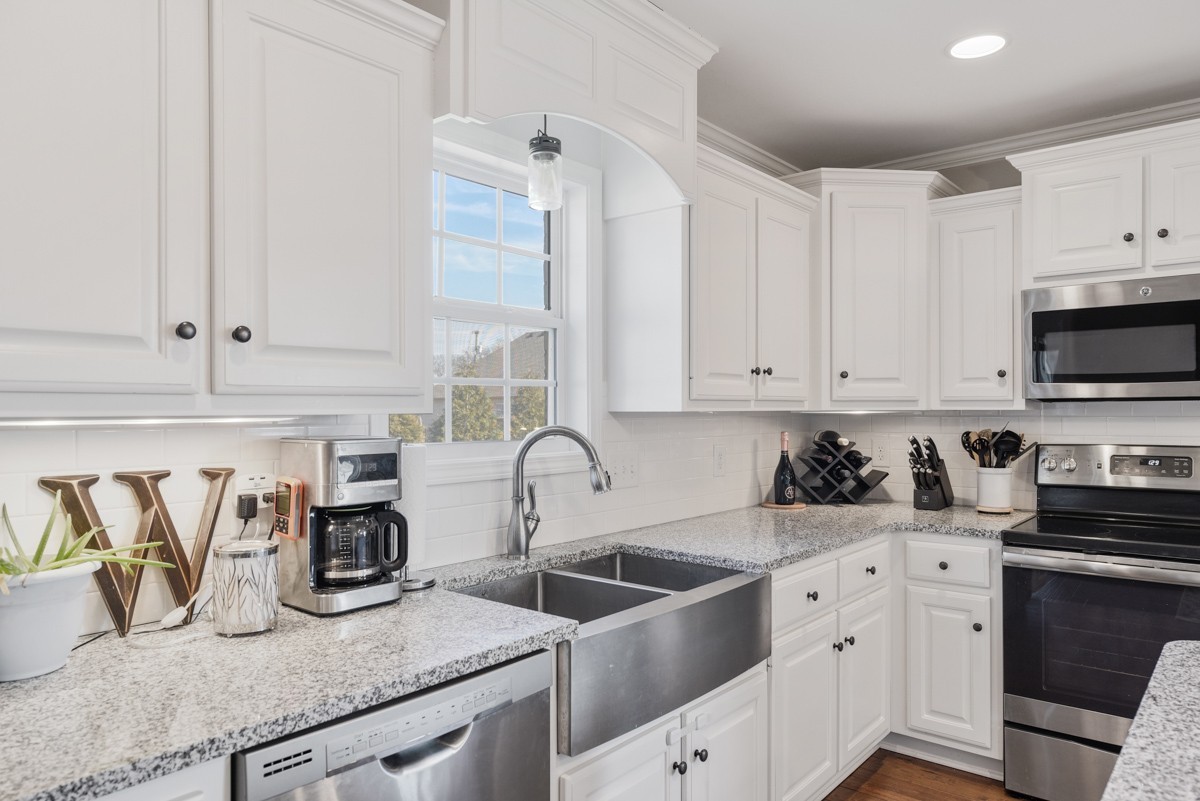 White kitchen with a farmhouse sink beneath a window, granite counters, and stainless-steel appliances.
