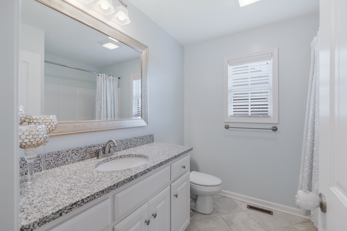 Bathroom with a granite vanity, large mirror, tiled floor, and window with blinds.
