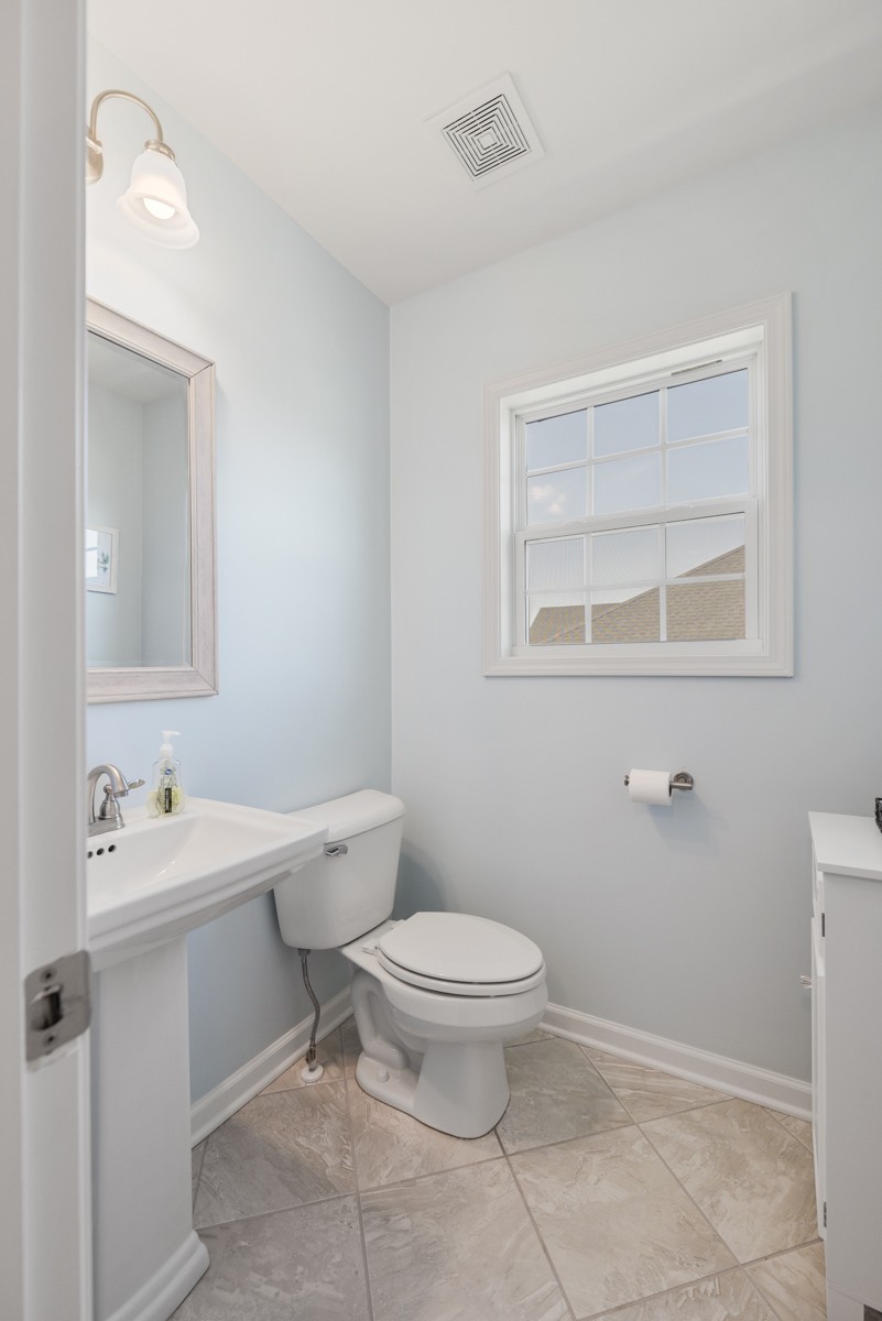 Bathroom with pale blue walls, a pedestal sink, white toilet, and window.