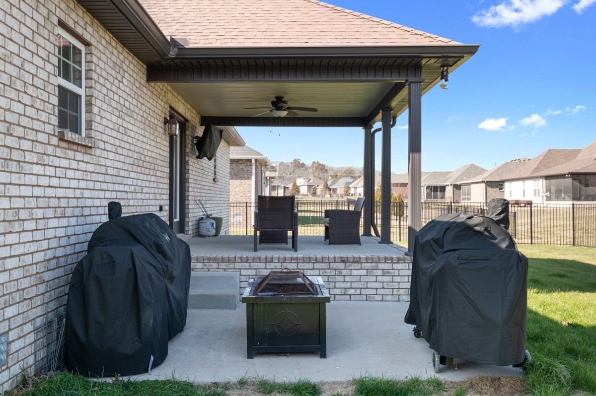 Covered brick patio with a ceiling fan, two chairs, covered grills, and a fire pit.