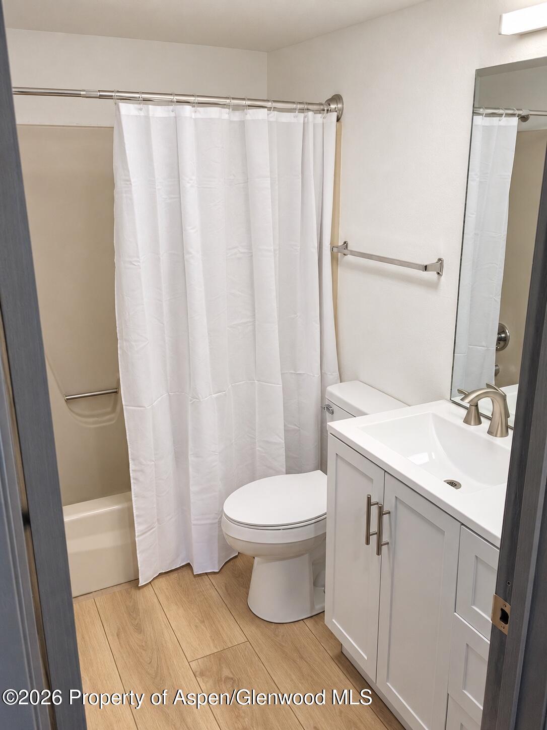 Bathroom with a curtained tub, white vanity, toilet, and light wood-look flooring.