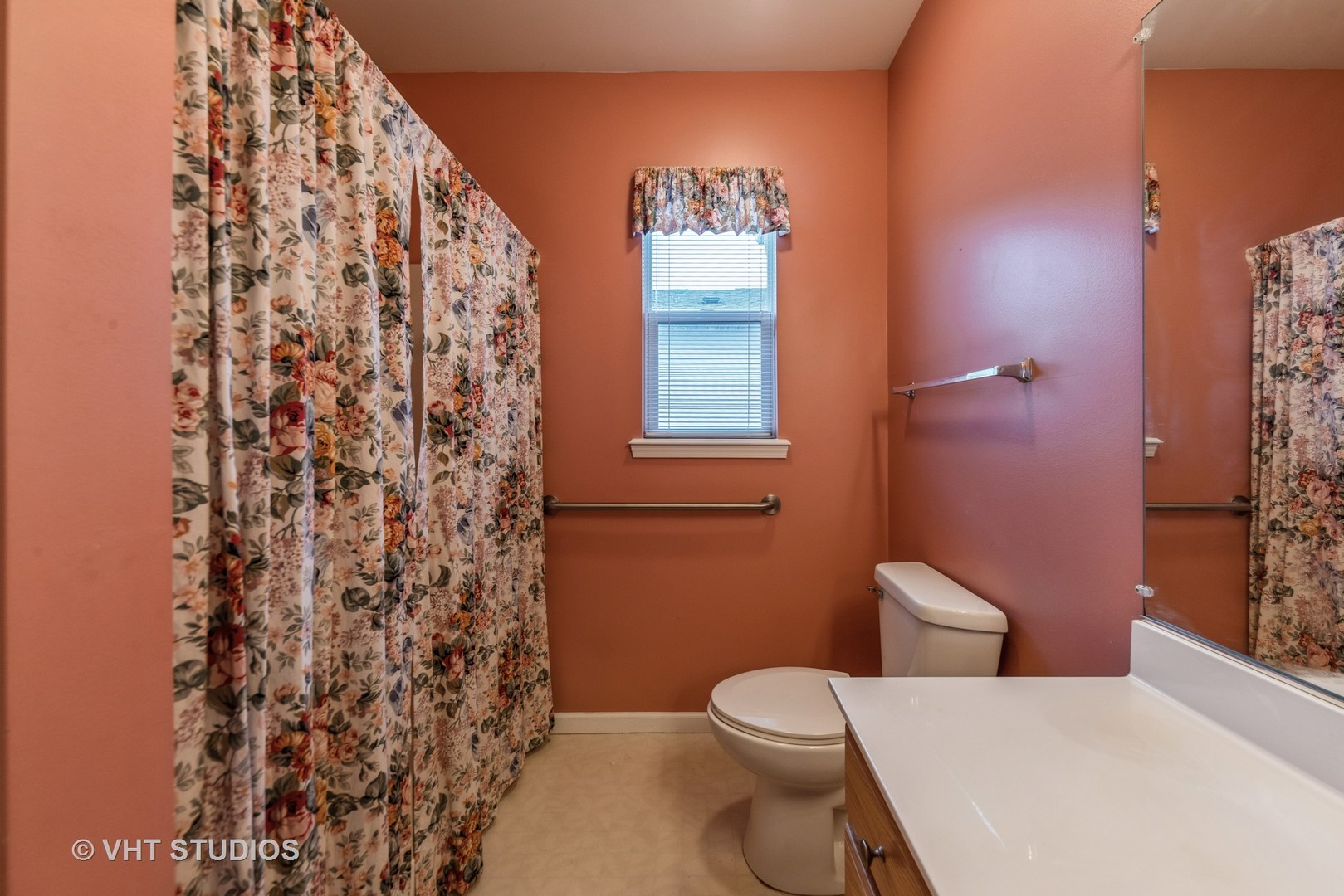 Bathroom with terracotta walls, floral shower curtain and window valance, and a large mirror.