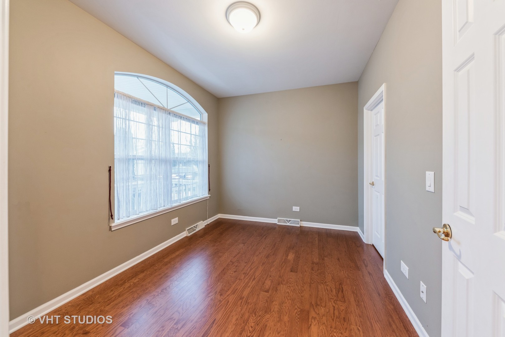 Empty room with hardwood floors and a large arched window with sheer curtains.
