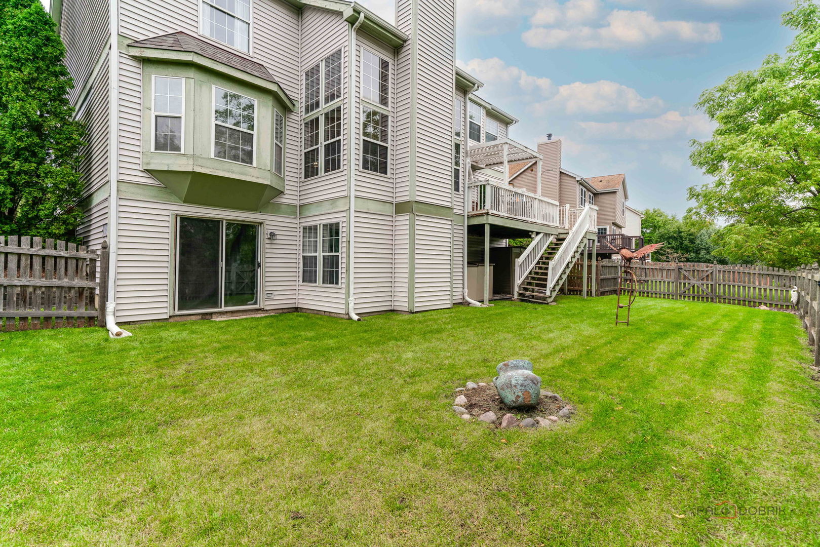 Home’s rear exterior with raised deck, stairs, and a fenced grassy backyard.