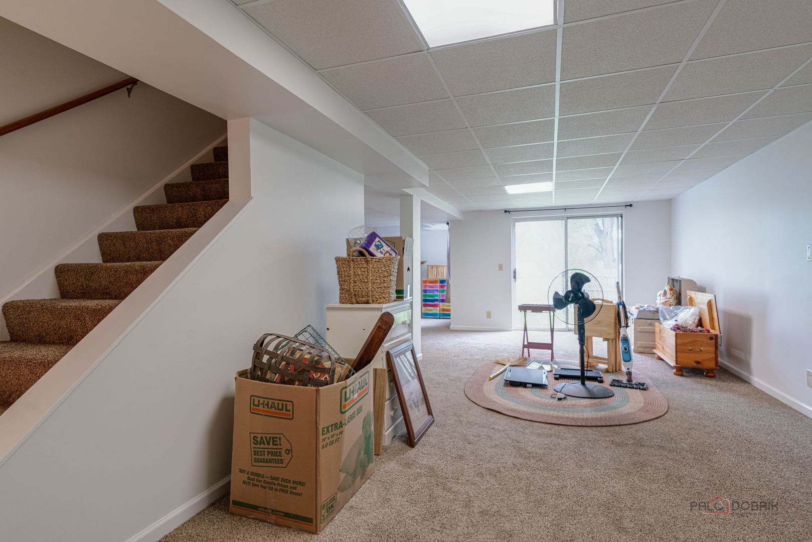 Carpeted basement with stairs, a sliding glass door, moving boxes, and stored belongings.