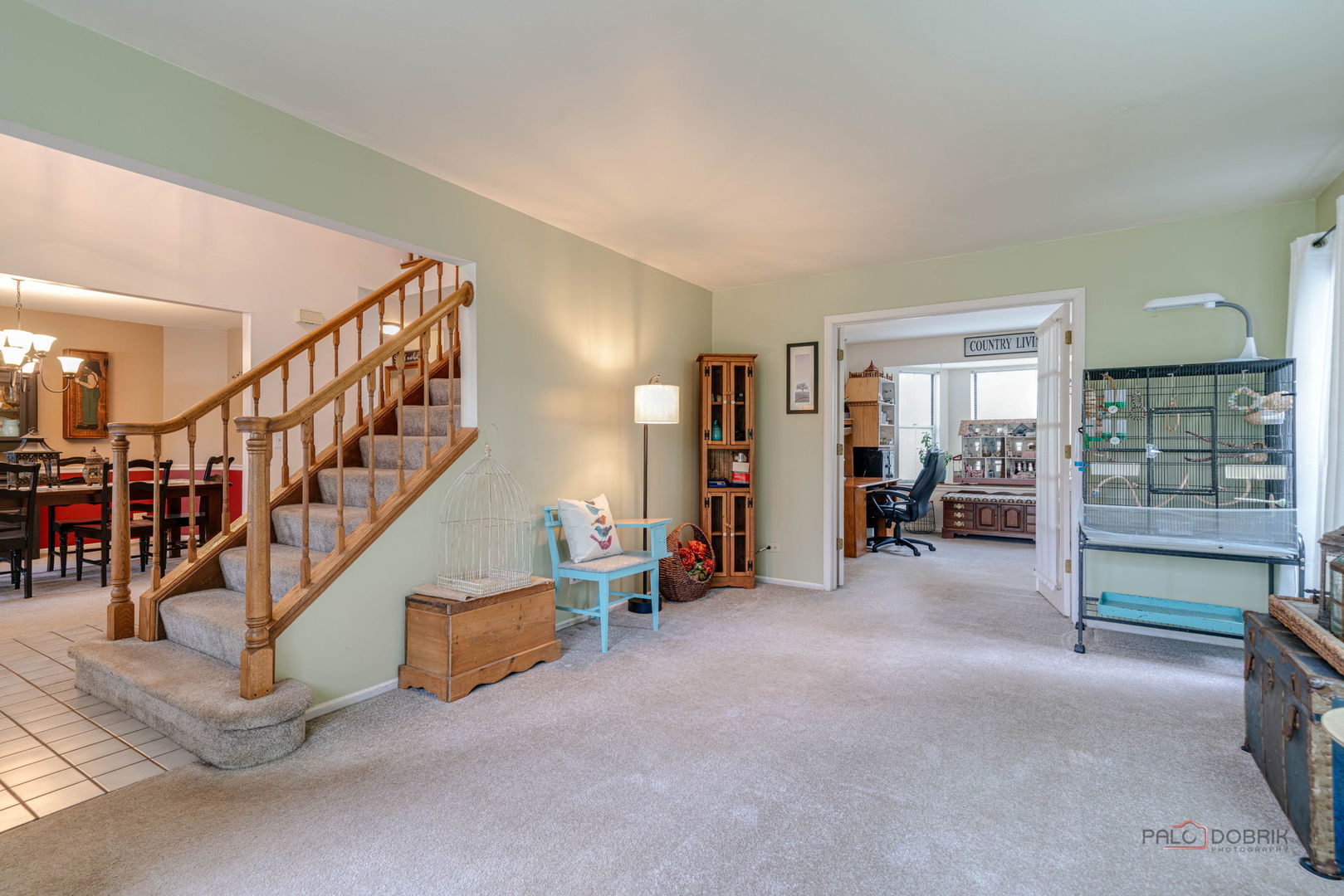 Carpeted living room with a wooden staircase and a home office visible through the doorway.