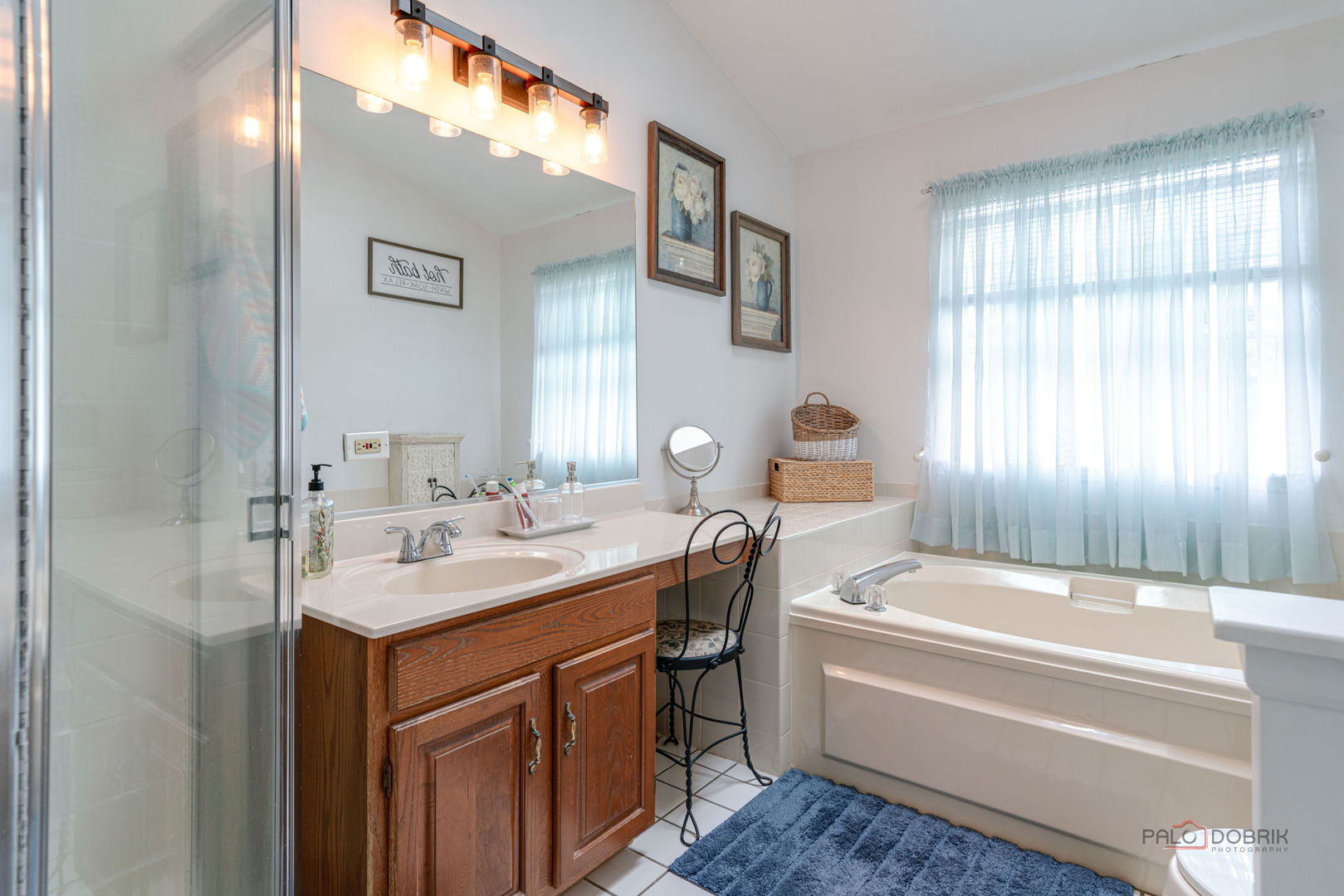 Bathroom with a wood vanity, glass shower, and built-in tub beneath a large curtained window.