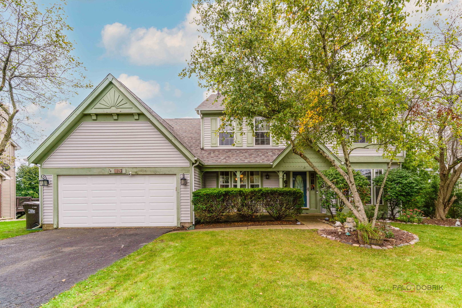 Two-story home with a double garage, green trim, mature trees, and a landscaped front lawn.