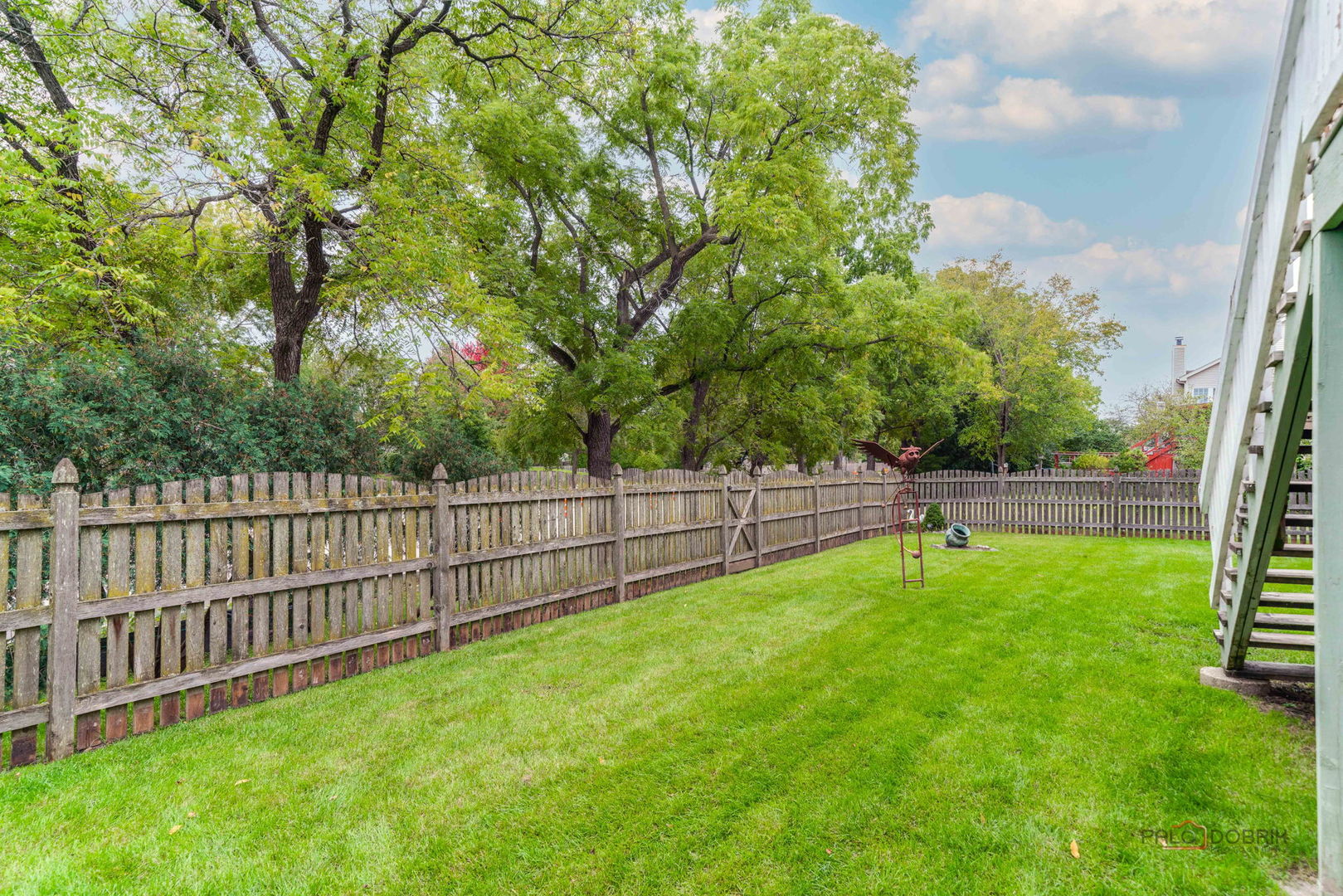 Fenced backyard with a broad lawn, mature trees, metal garden sculpture, and exterior stairs.