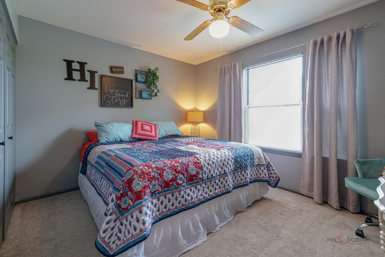 Bedroom with a colorful patterned quilt, ceiling fan, and bright curtained window.
