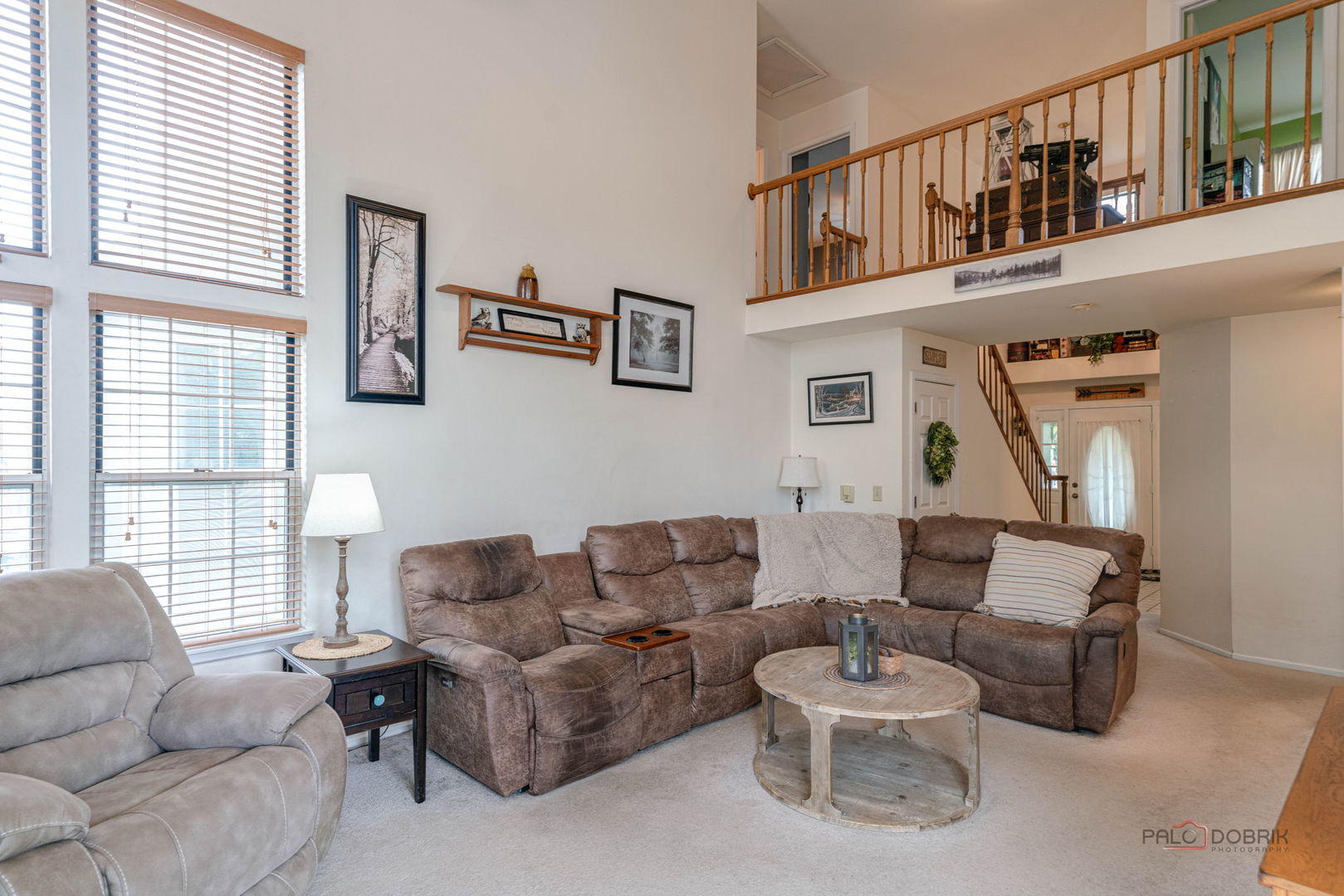 Living room with a brown sectional, tall windows, and a wooden-railed upper-level landing.